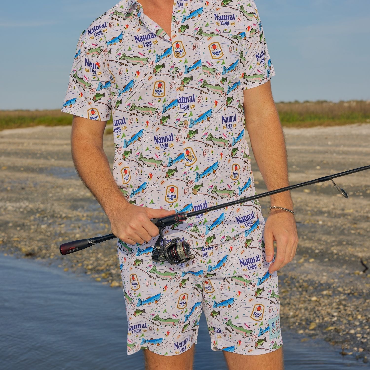 Person wearing a patterned outfit with 'Natural Light' branding, holding a fishing rod on a beach.