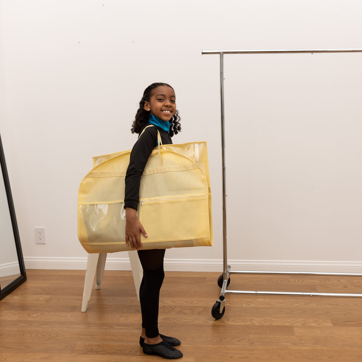Person holding a yellow garment bag in a room with a white wall and wooden floor.