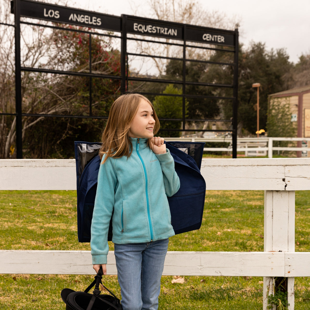 Girl in teal jacket standing next to a blue bag at the Los Angeles Equestrian Center.