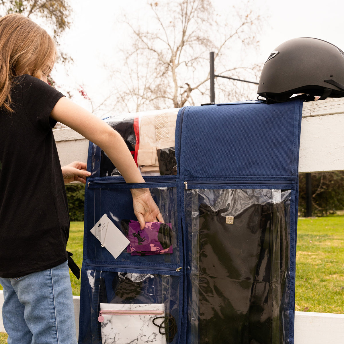 A navy blue fabric hanging organizer with clear pockets is shown outdoors next to a white fence, with a person reaching into one of the pockets and a black helmet resting on top.