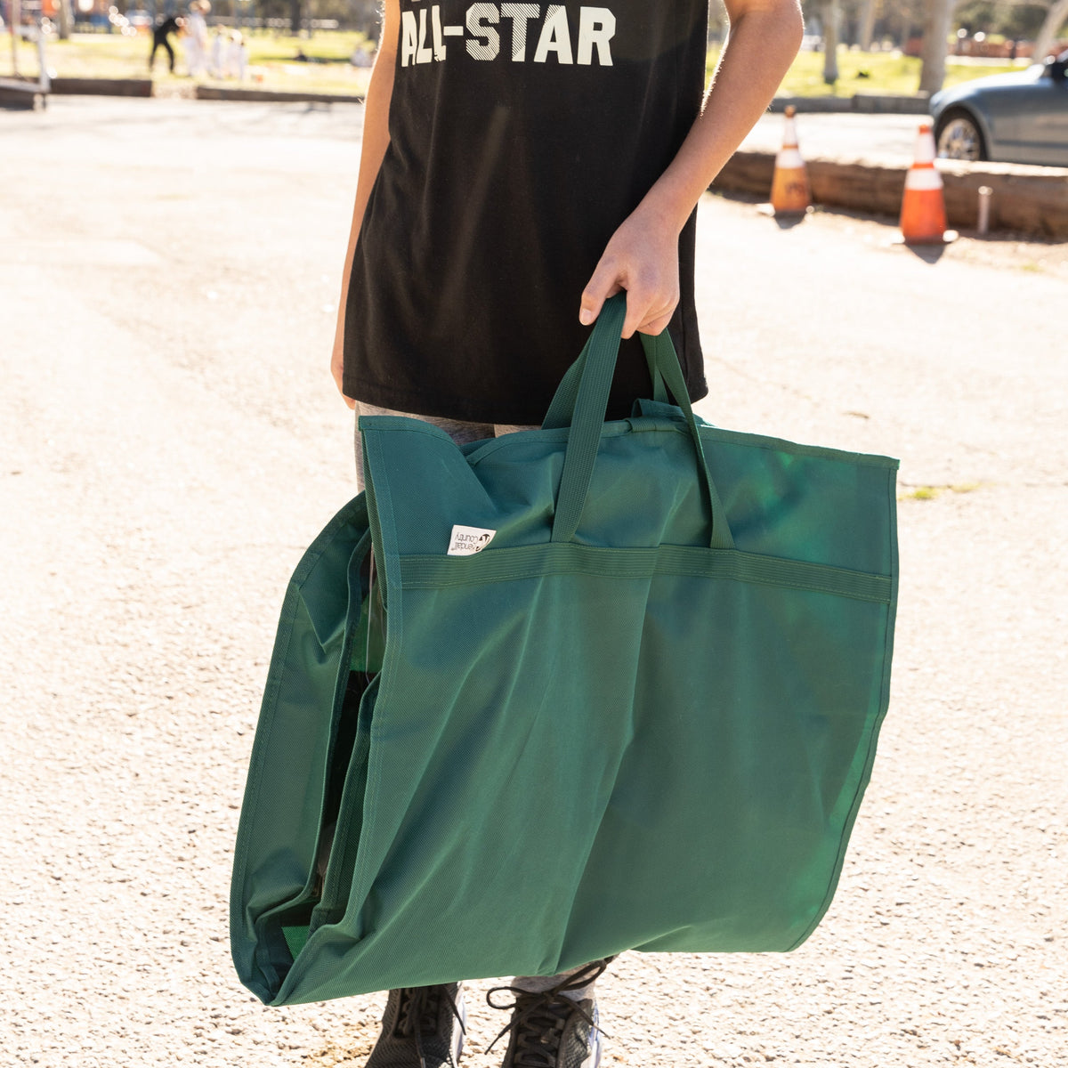 A person outdoors carrying a large dark green reusable tote bag with two handles. The person is wearing a black t-shirt with 'ALL-STAR' printed in white.