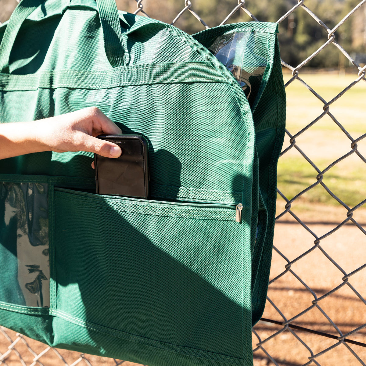 A green canvas car window organizer hanging on a chain-link fence, with a hand placing a black wallet into one of its front pockets. The background shows a blurred outdoor baseball field.