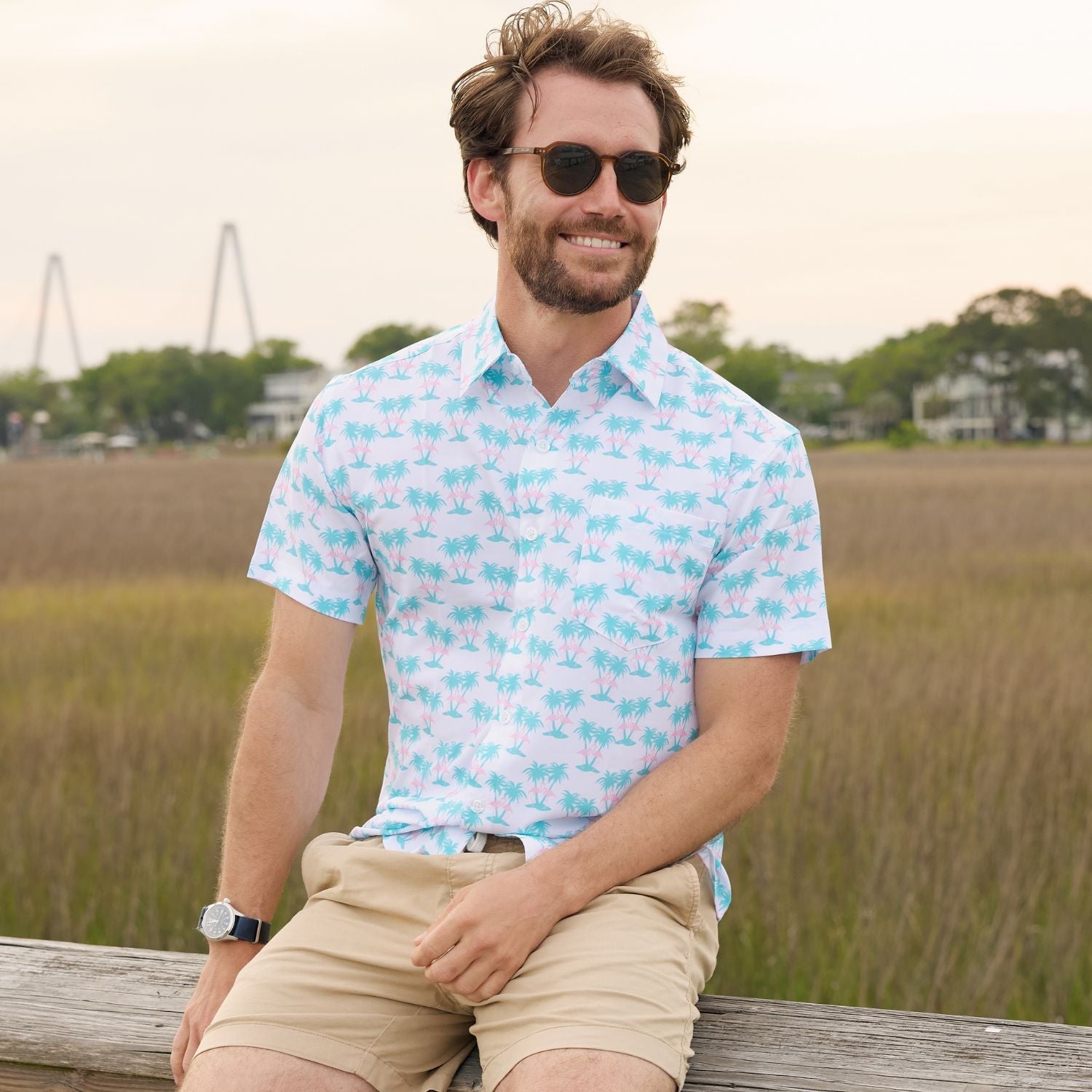 Man wearing a light blue patterned shirt and sunglasses sitting outdoors.