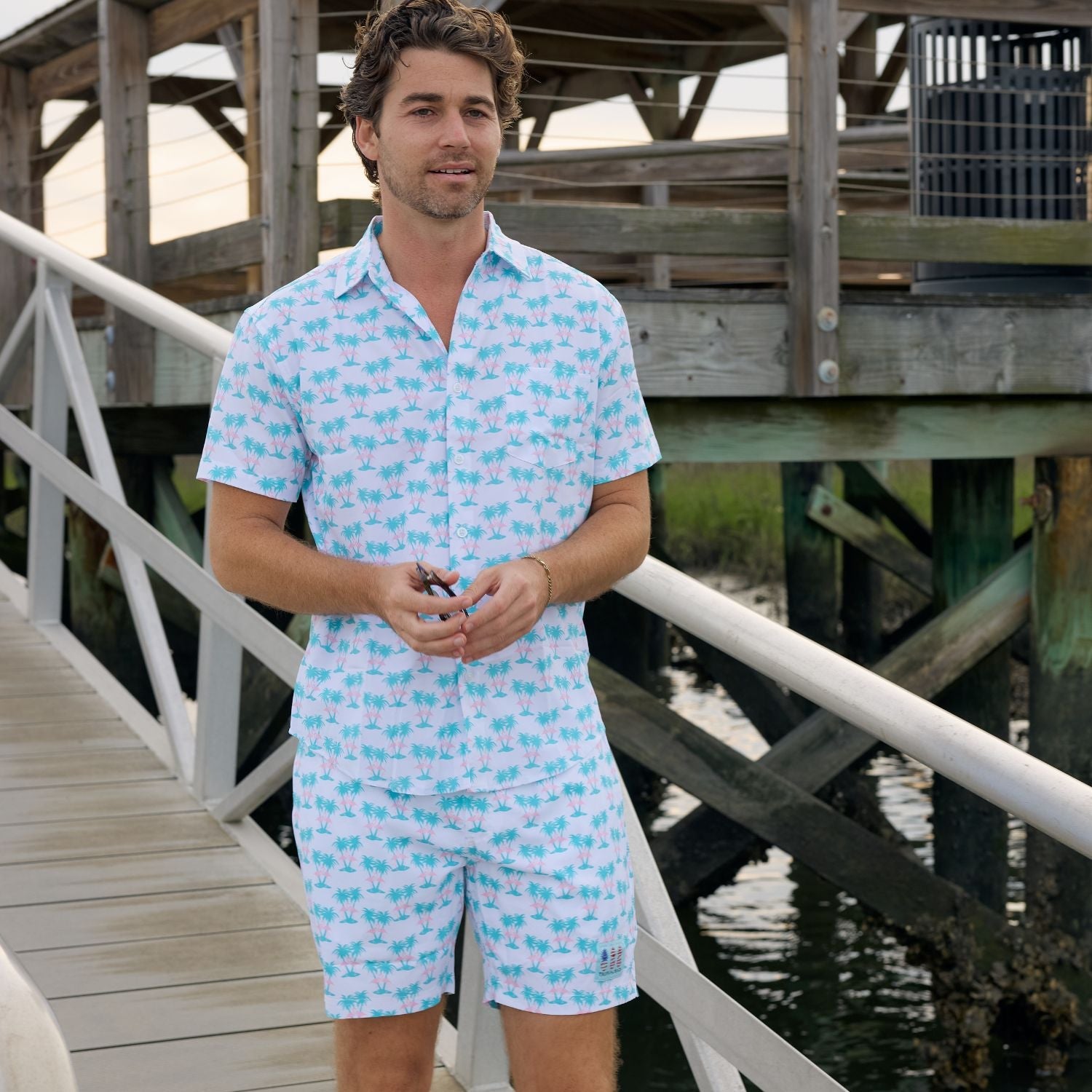 Man wearing a light blue patterned romper standing on a wooden dock by water.