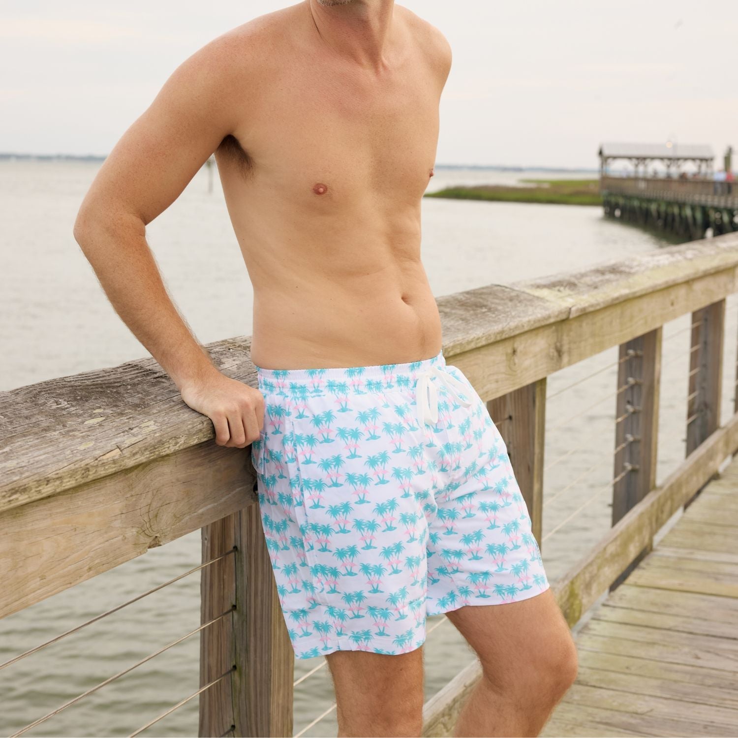 Man wearing light blue swim trunks with a pattern standing on a wooden dock by a body of water.