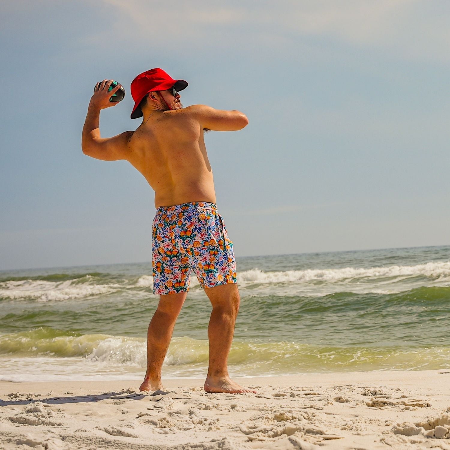 Man in colorful swim trunks and red cap standing on a beach, taking a selfie.