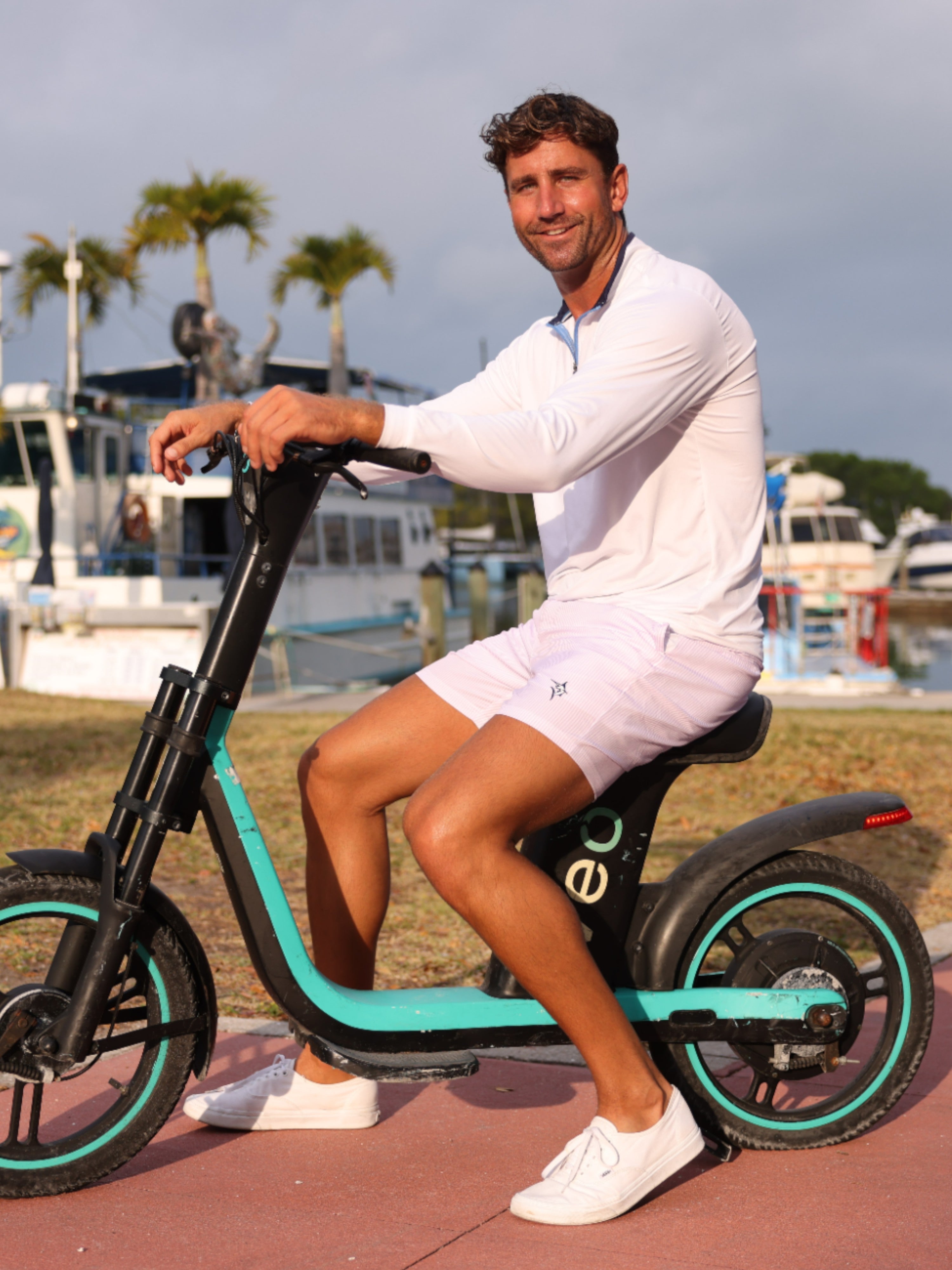Man riding a teal and black scooter by a waterfront with palm trees in the background
