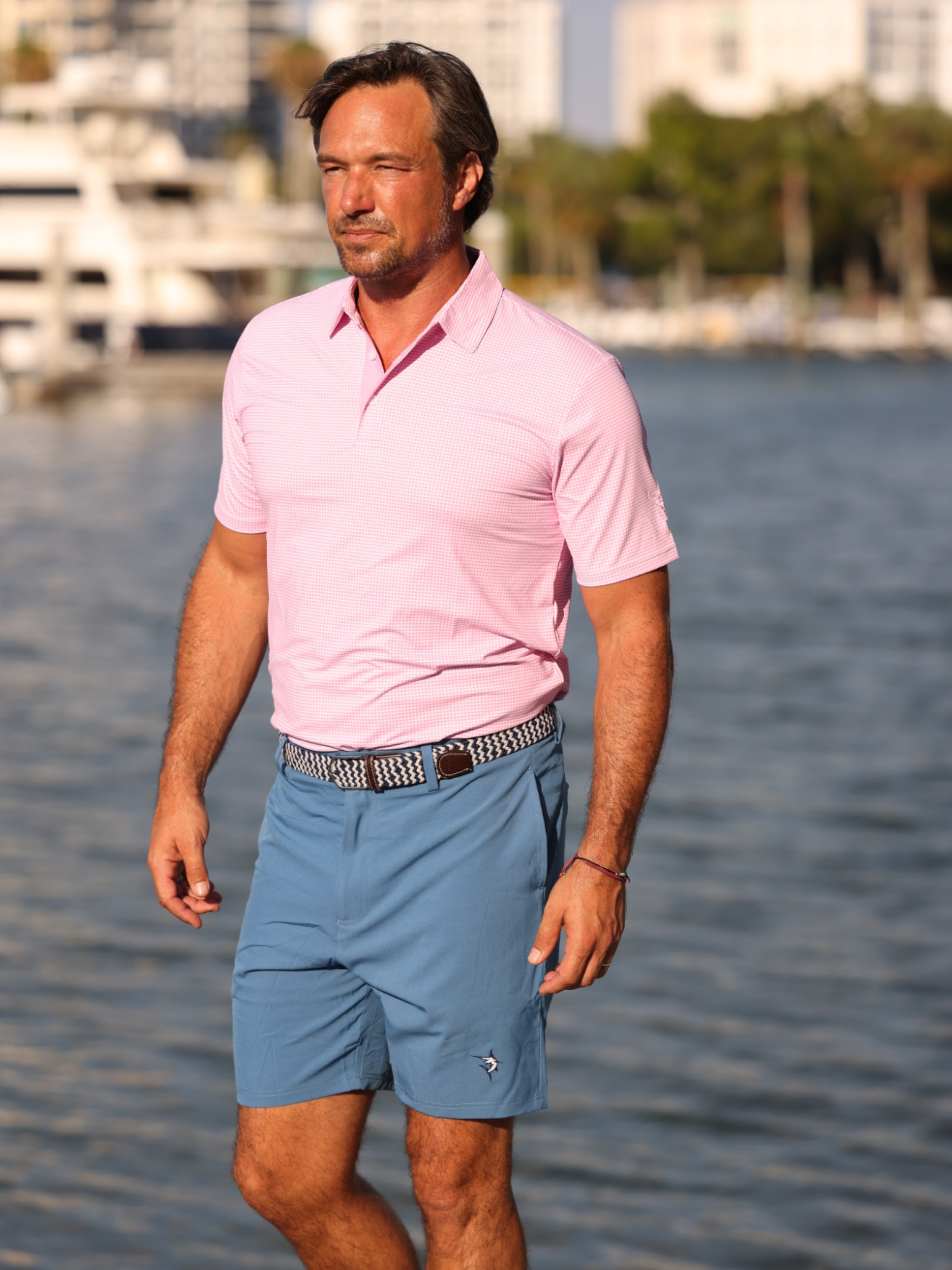 Man wearing a pink polo shirt and blue shorts standing by a waterfront with buildings and trees in the background.