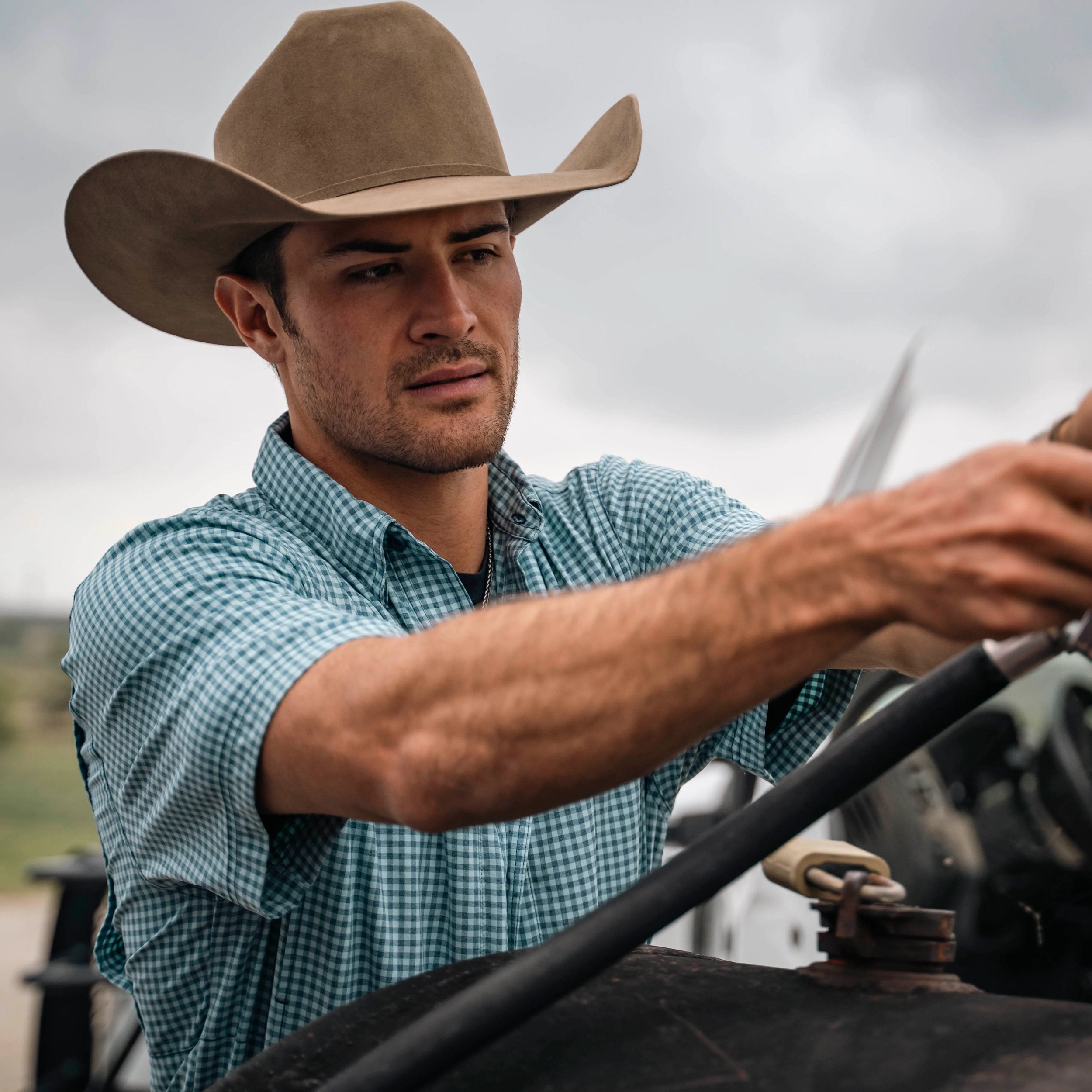 Man wearing a cowboy hat and checkered shirt, holding onto a vehicle's steering wheel.