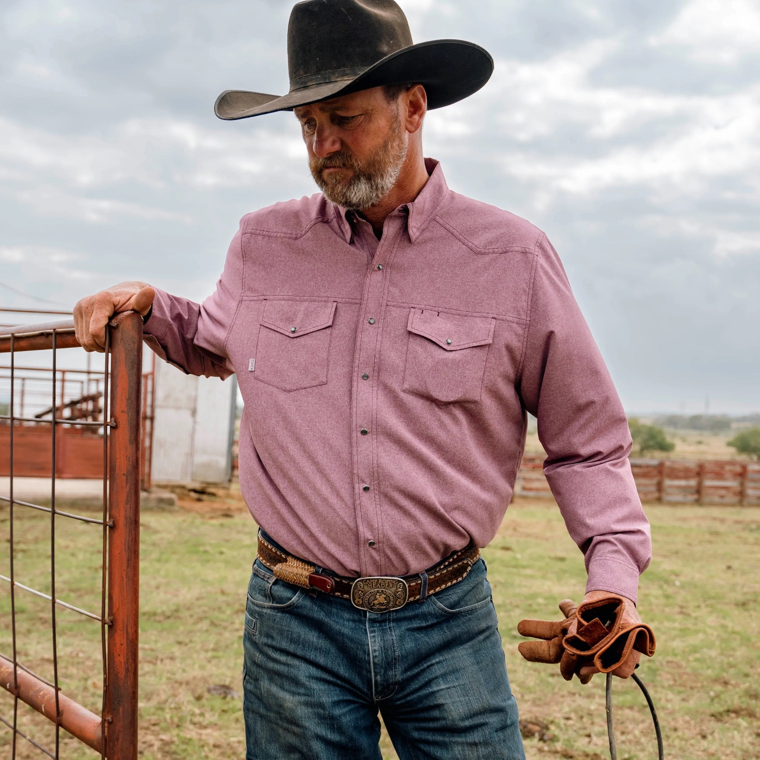 Man in cowboy attire standing in a field with a cloudy sky.