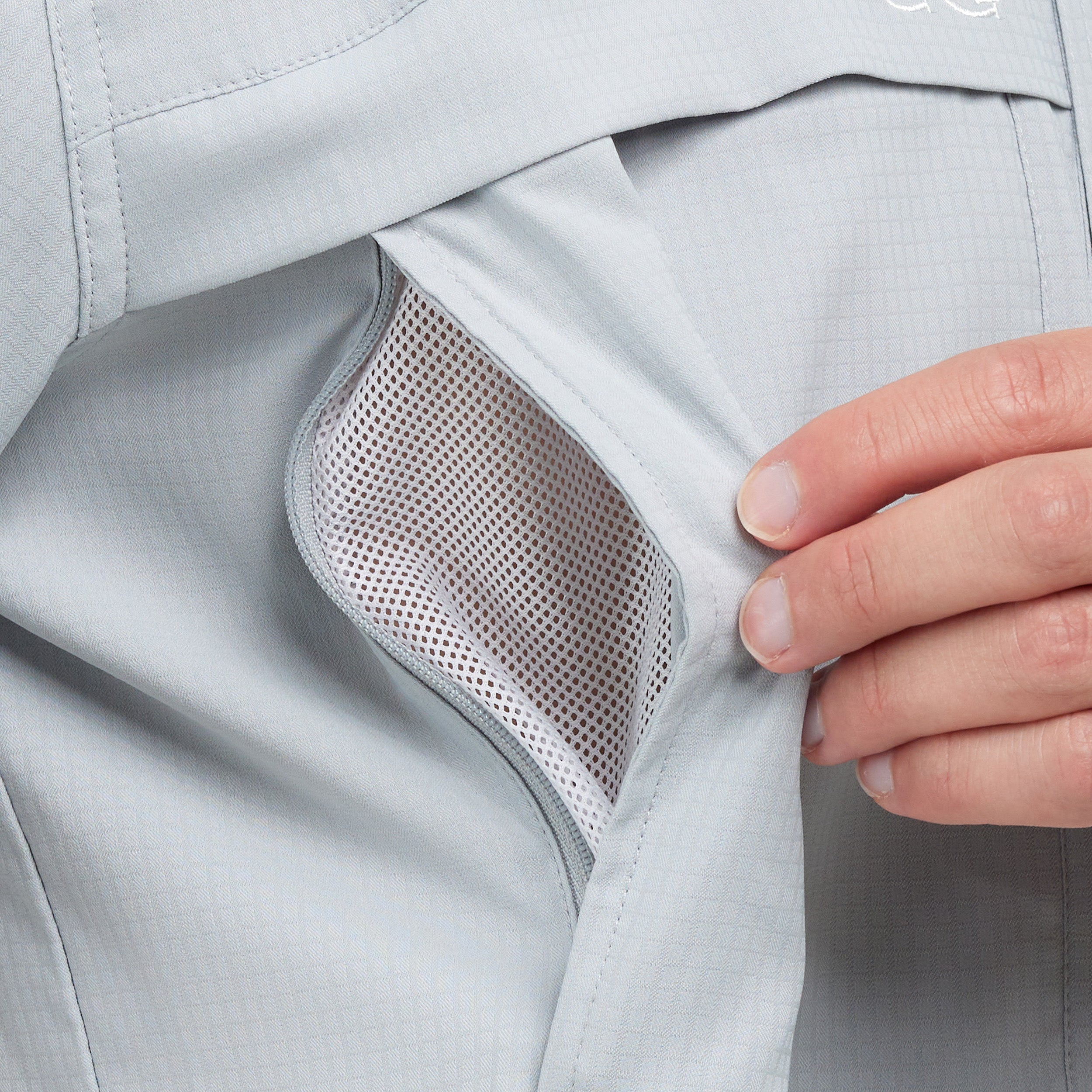 Close-up of a light gray technical jacket with a hand opening a zippered pocket to show the breathable mesh lining.