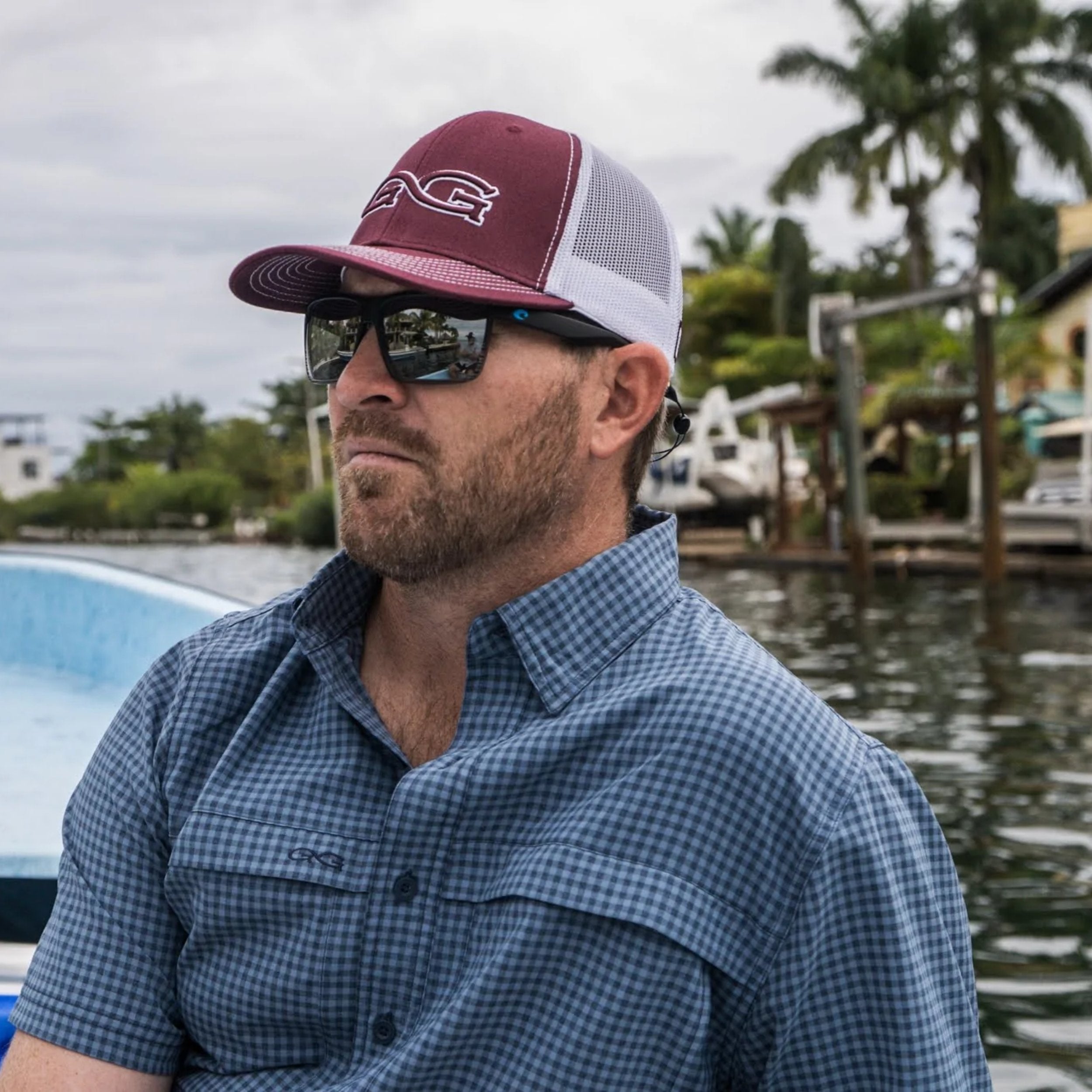 Man wearing a maroon and white cap with sunglasses and a checkered shirt by a waterfront.