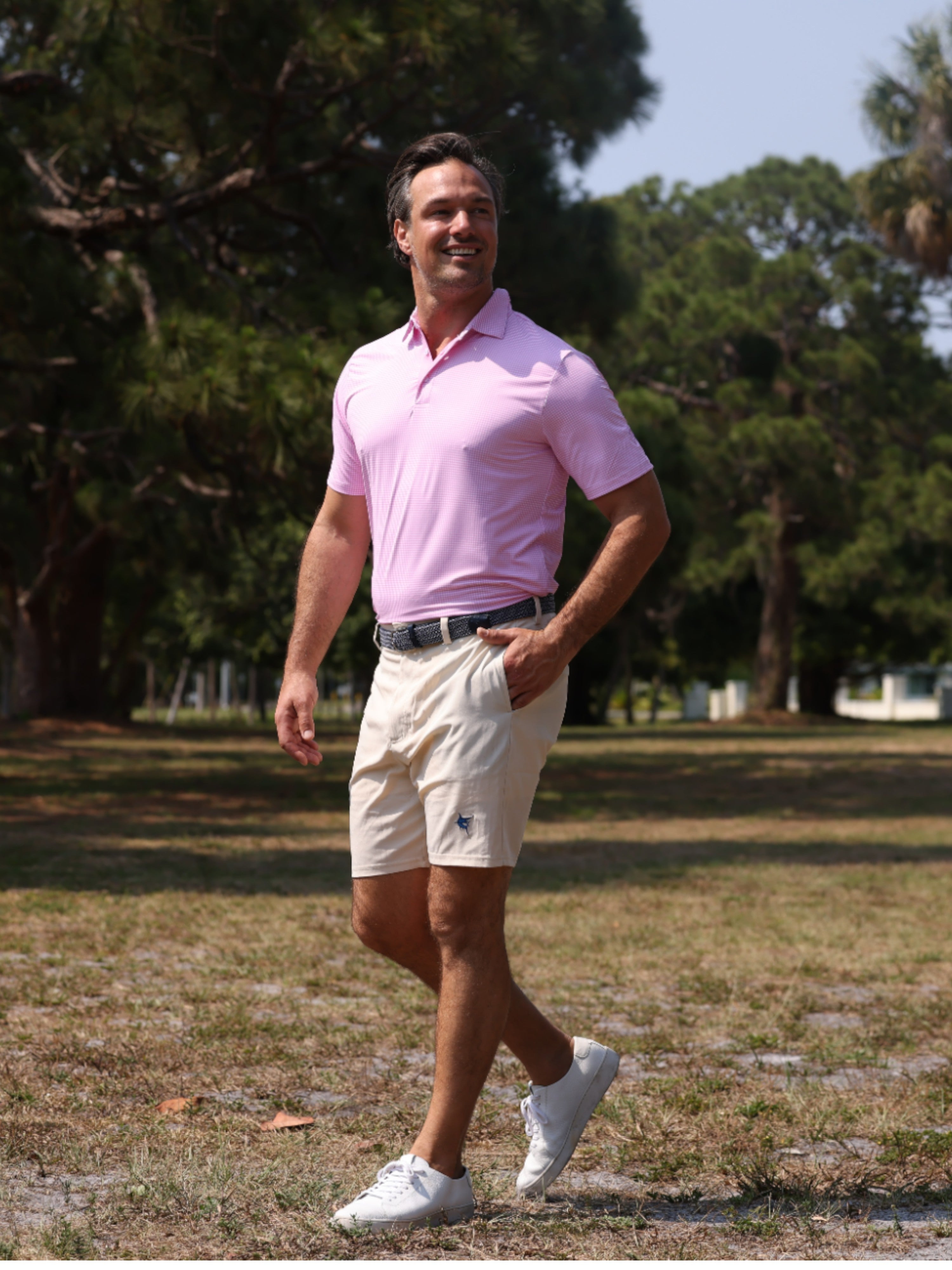 Man in a pink polo shirt and beige shorts walking outdoors with trees in the background