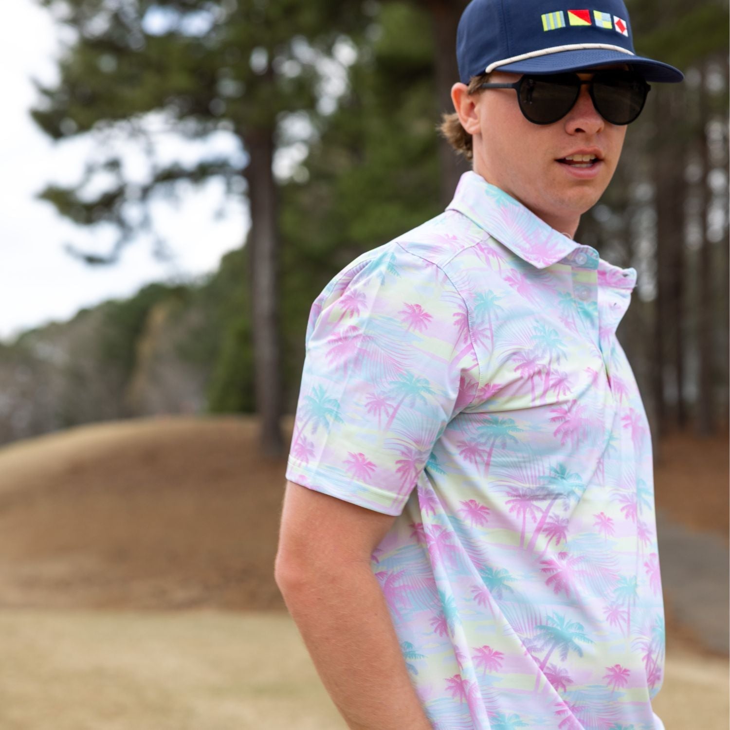 Man wearing a colorful patterned shirt and navy cap with flags, standing outdoors.