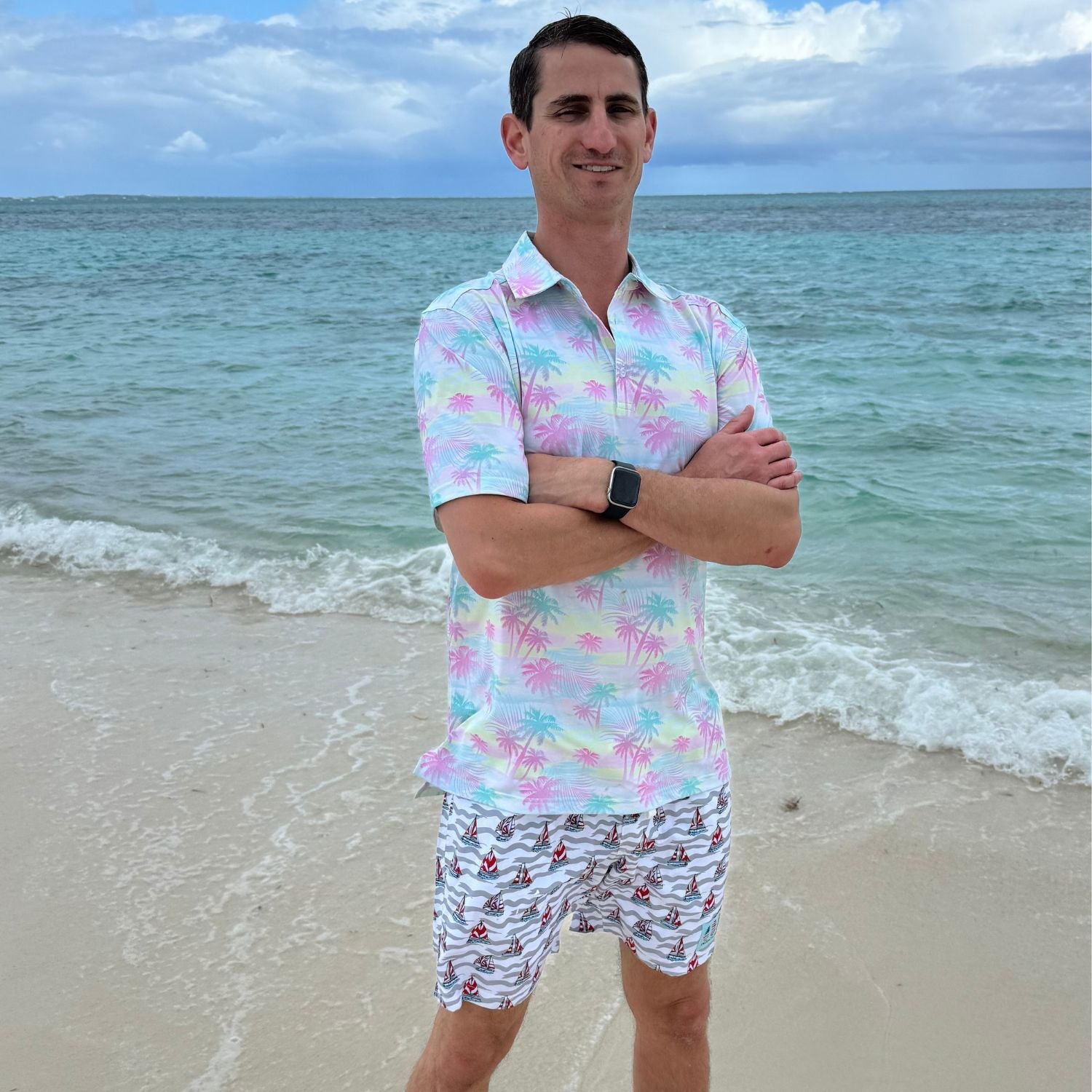 Man standing on a beach wearing a colorful shirt and patterned shorts with ocean in the background