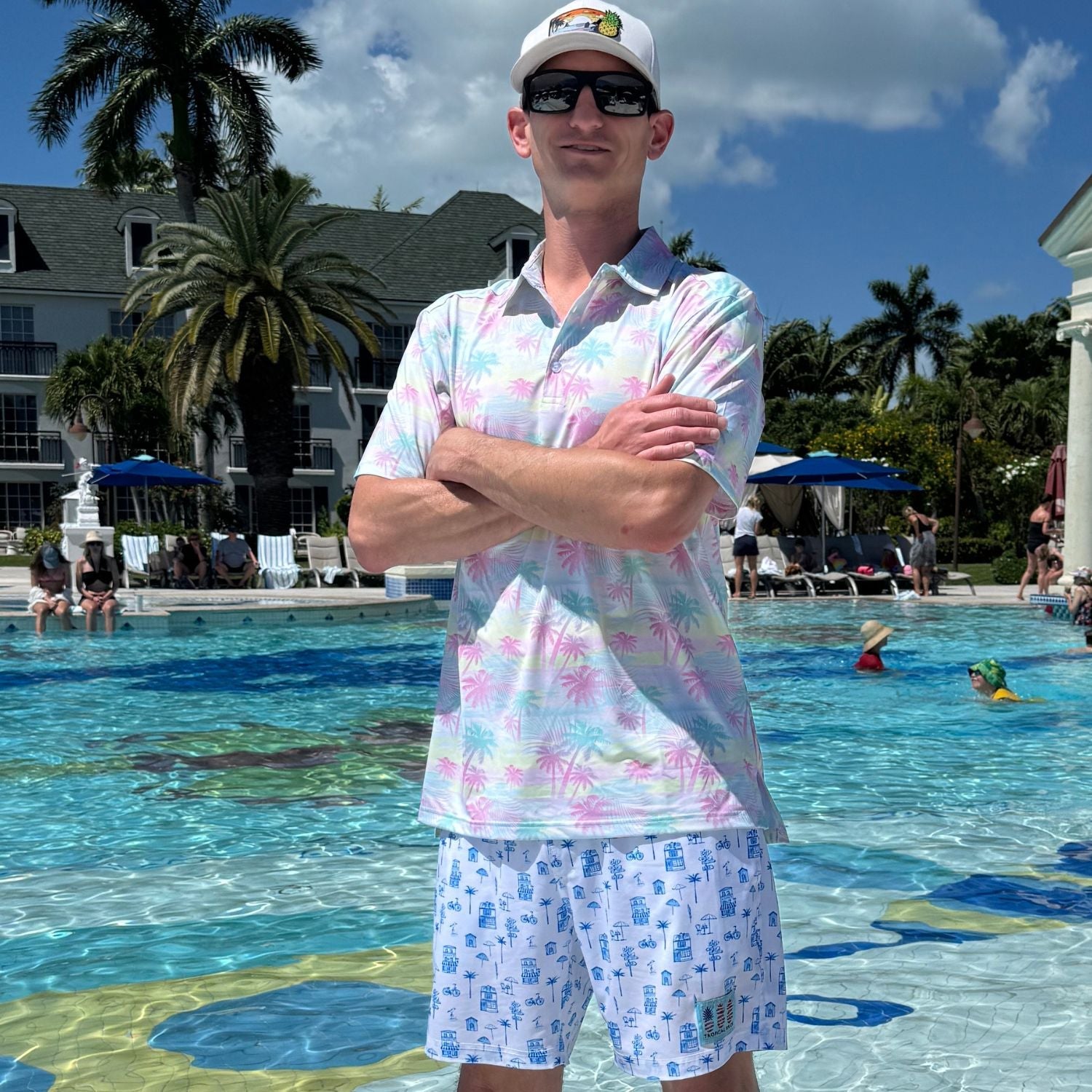 Man in a colorful shirt and shorts standing by a pool with palm trees and people in the background.