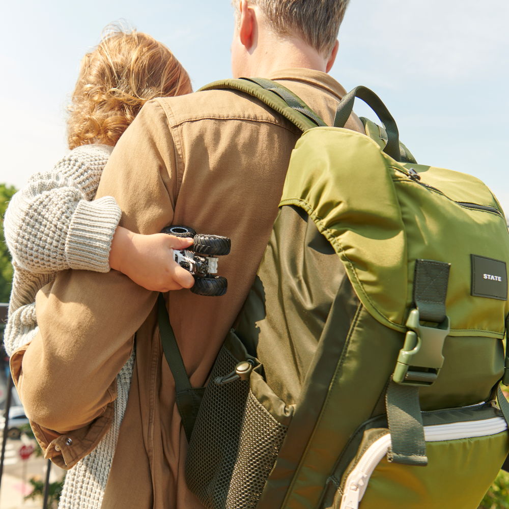 Person wearing a green backpack with a child holding a camera, outdoors.