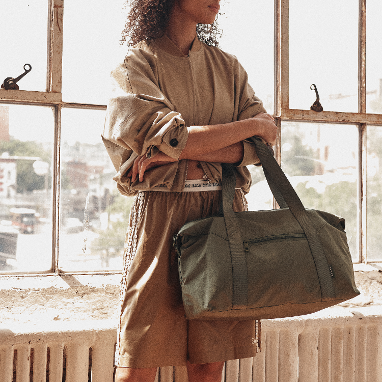 A person standing by a sunlit window holds a large olive green duffel bag. The person is wearing a beige cropped sweatshirt and matching shorts.