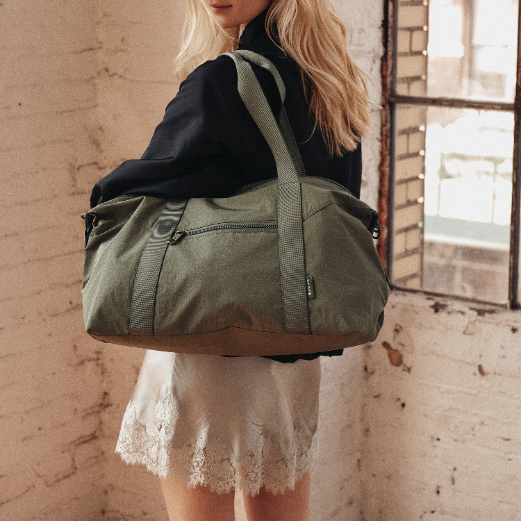 A woman wearing a black jacket and a white lace hem dress carries an olive green duffel bag over her shoulder in a rustic room with a brick wall and window.
