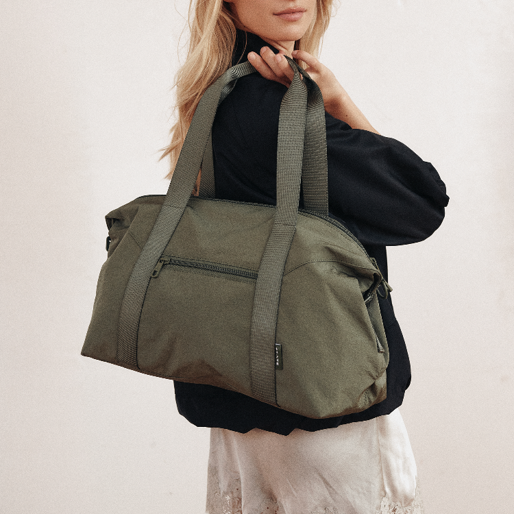 An olive green duffel bag with wide shoulder straps, shown being carried over the shoulder of a model wearing a black top and white skirt against a plain off-white background.
