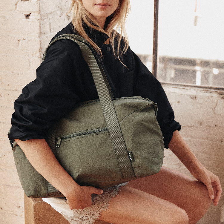 A woman wearing a black shirt and white skirt sits indoors holding an olive green duffel bag with a wide shoulder strap. The bag has a zippered front pocket and a small black brand tag.