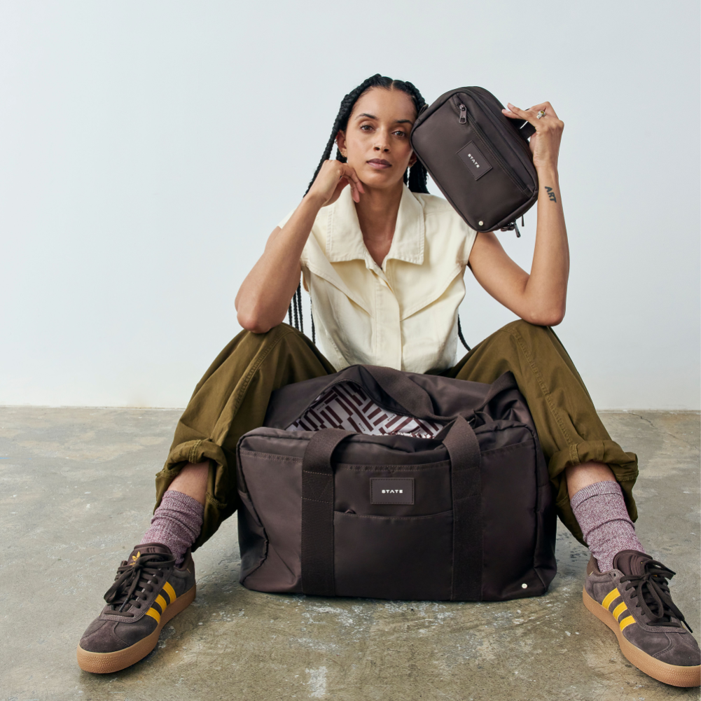 Woman sitting on the floor with a large duffel bag and a smaller bag, wearing a white shirt and green pants.
