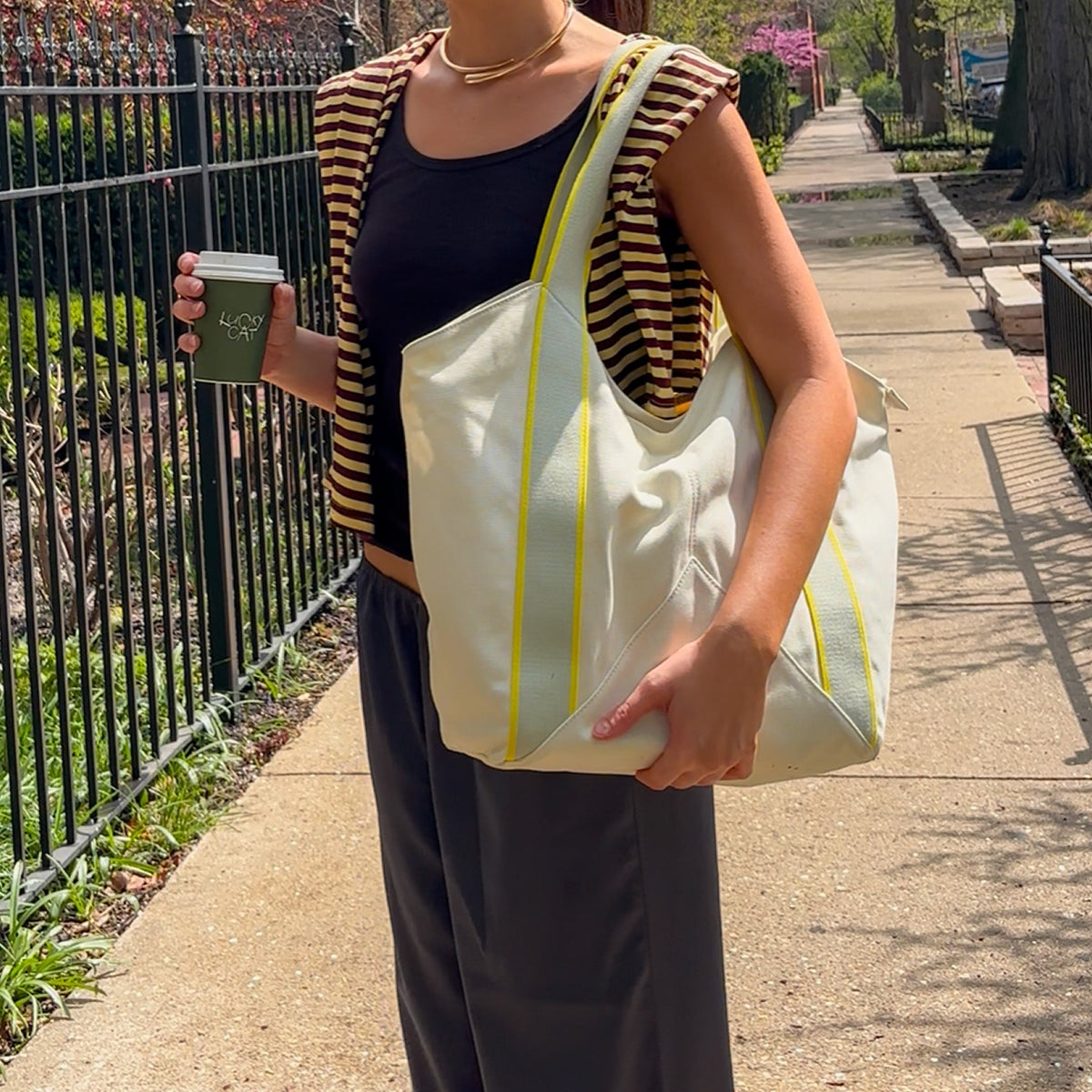 Person holding a white tote bag with yellow straps and a coffee cup outdoors.