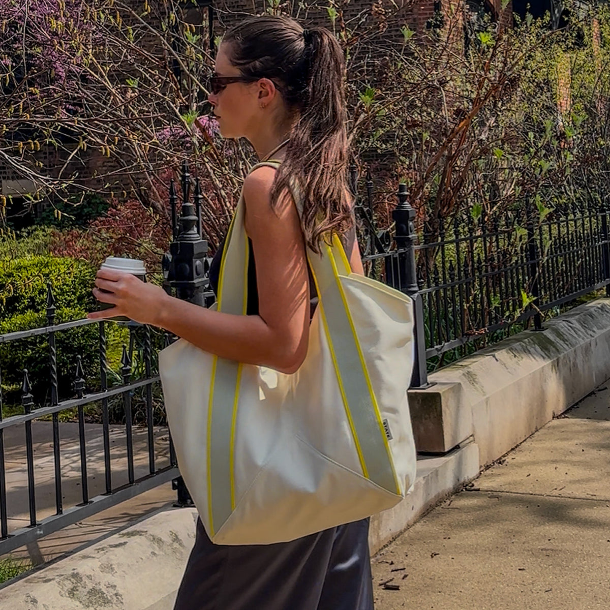 Woman holding a white tote bag with yellow accents outdoors.