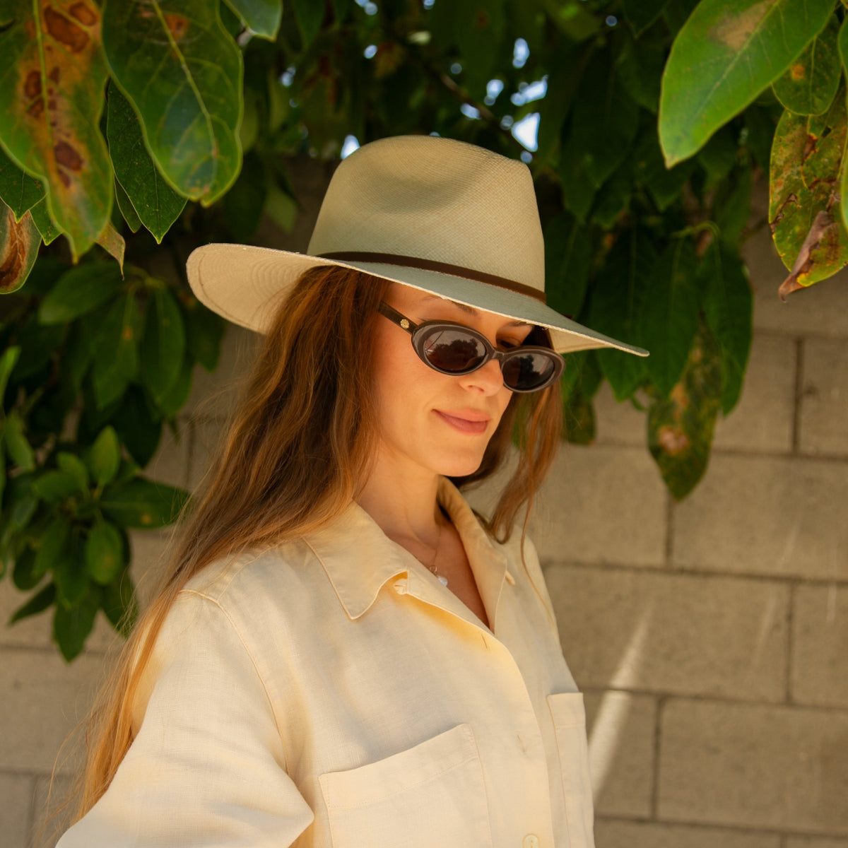 Woman wearing a beige hat and sunglasses against a leafy background