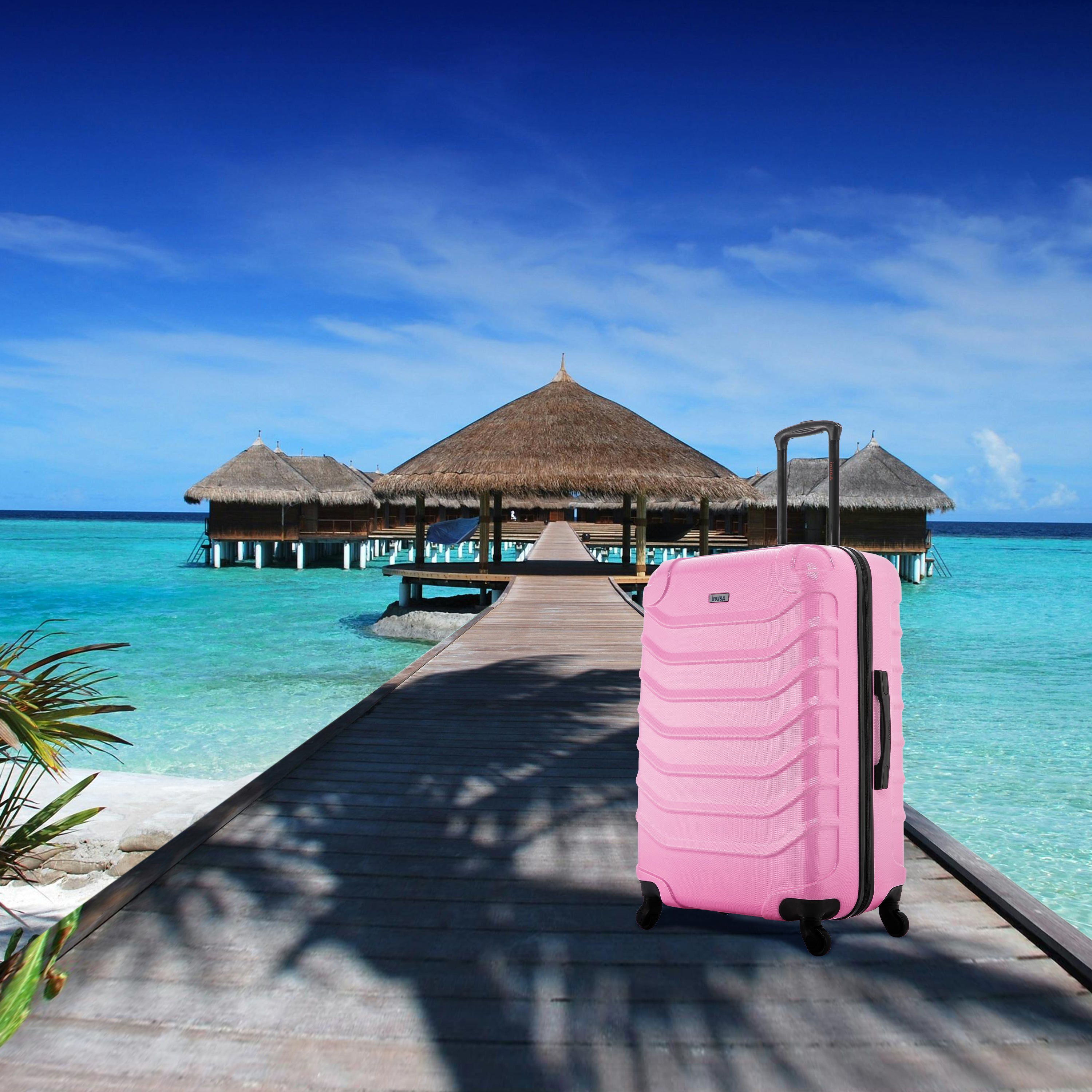 Pink suitcase on a wooden pier leading to thatched-roof huts over turquoise water.