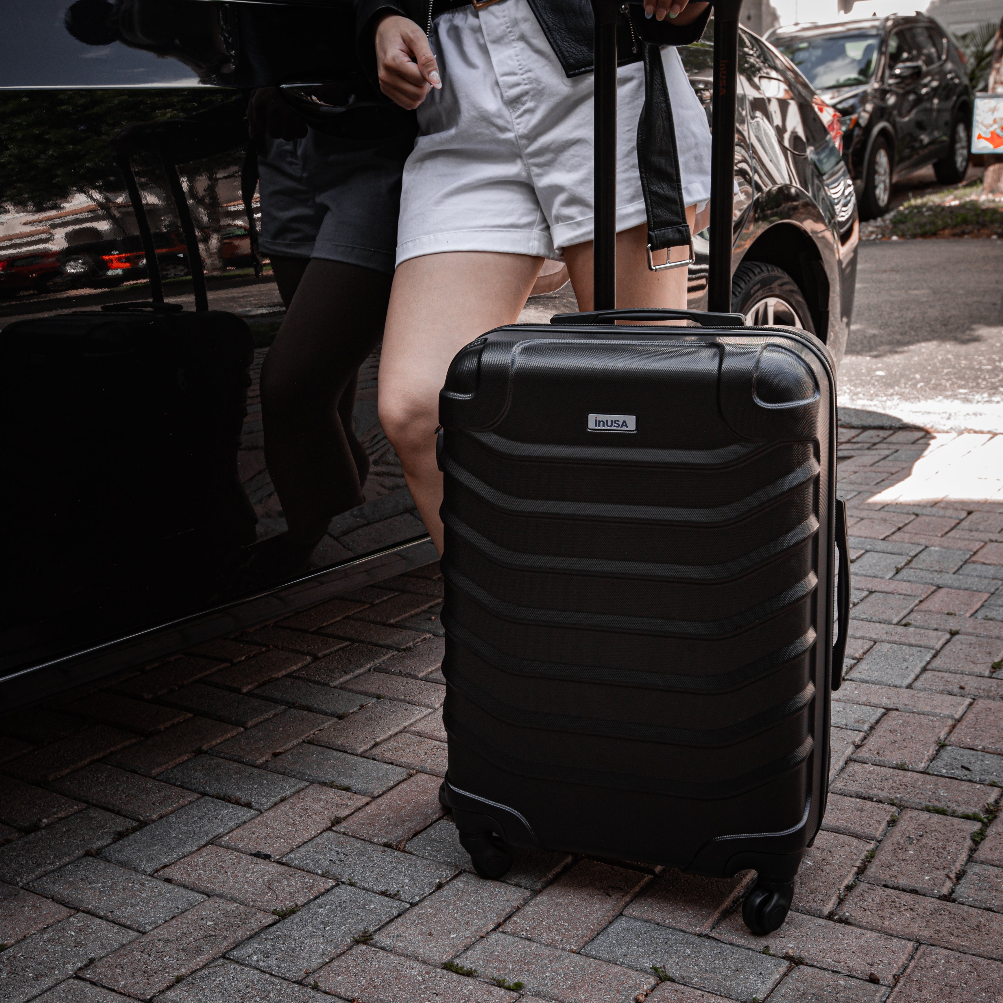 Person pulling a black suitcase on a sidewalk with cars in the background