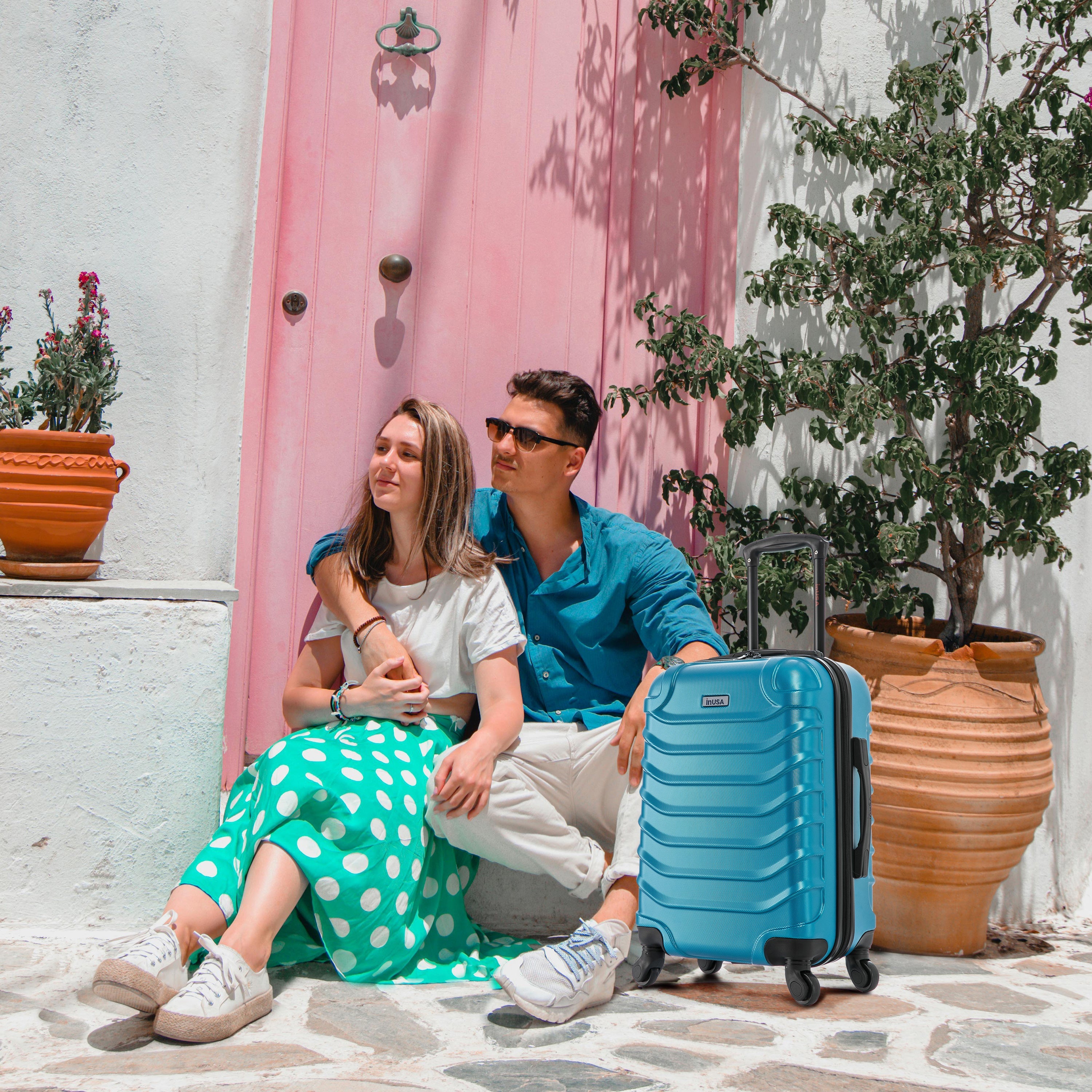 Couple sitting on a stone floor with a blue suitcase next to them, pink curtain and plants in the background.