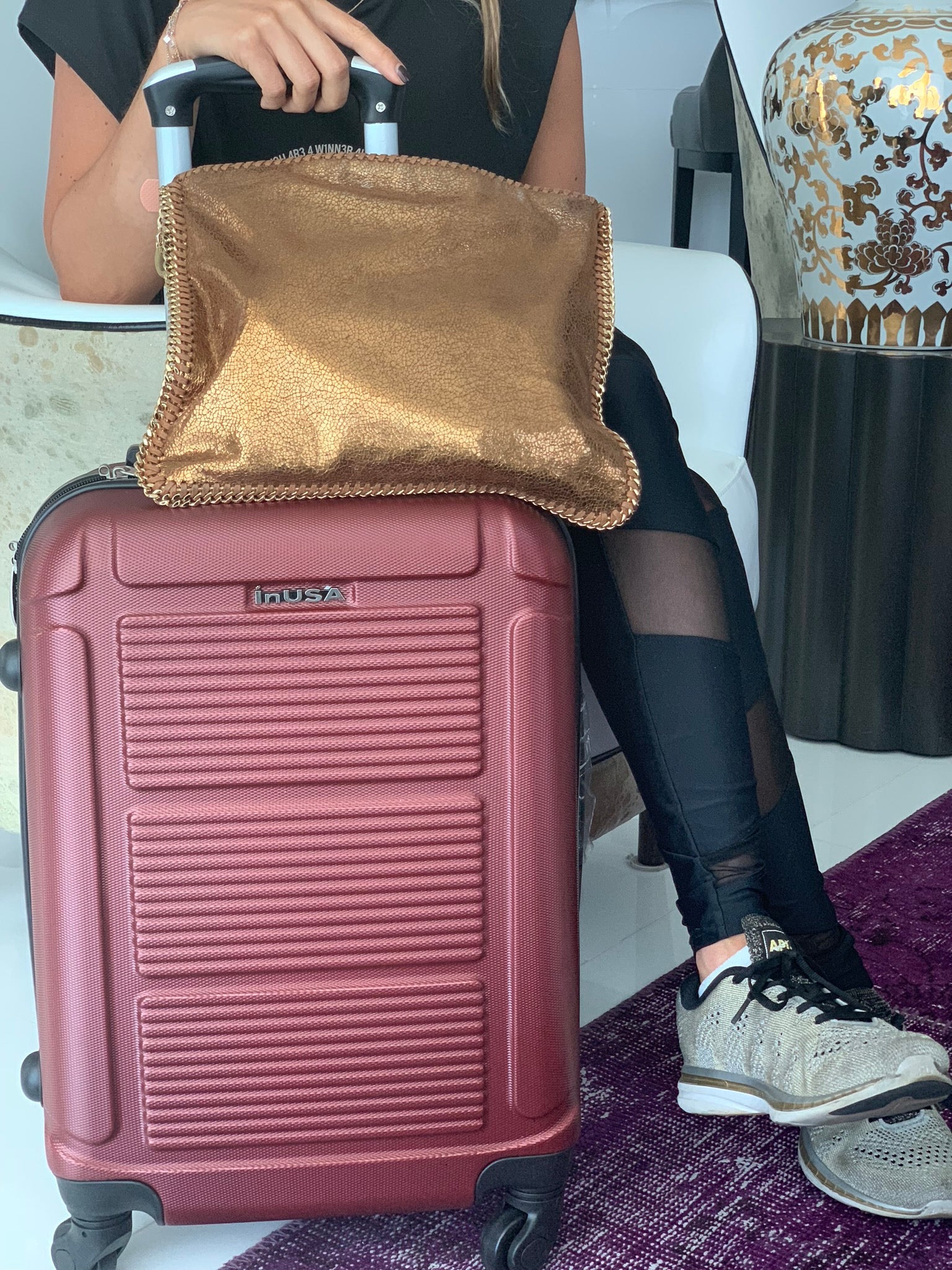 Person sitting on a suitcase with a gold bag on top, wearing black leggings and sneakers.