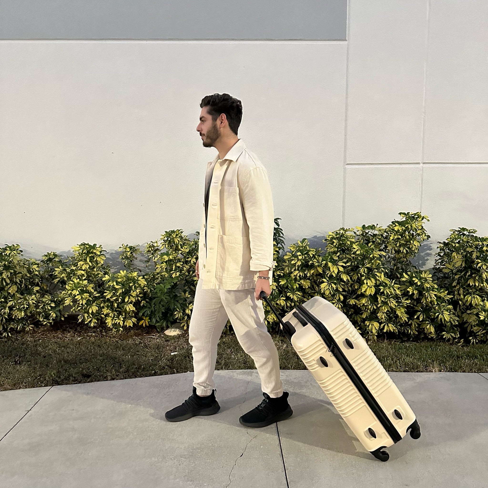 Man pulling a beige suitcase on a sidewalk with greenery in the background