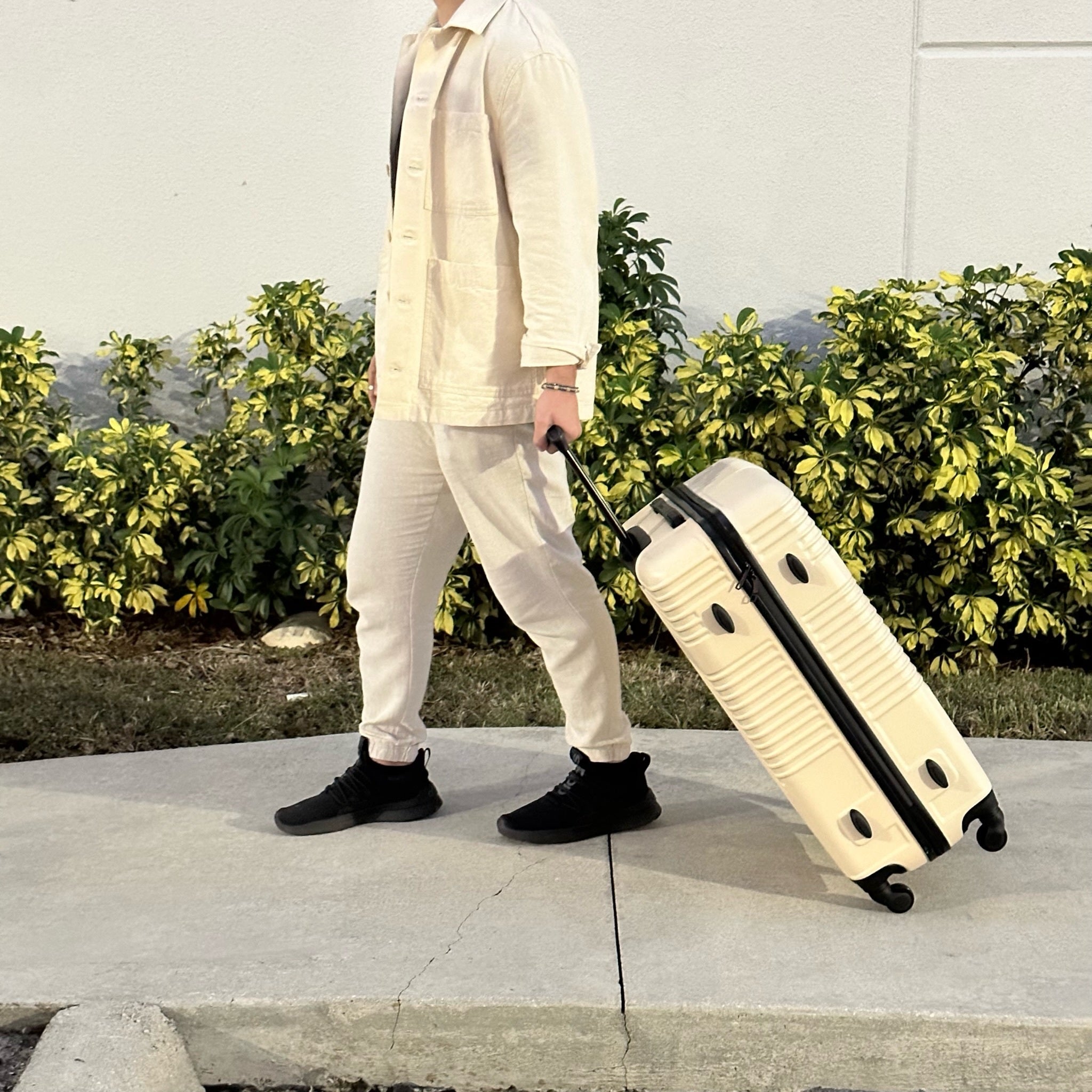 Person in beige jacket and pants pulling a cream suitcase on a sidewalk with greenery in the background