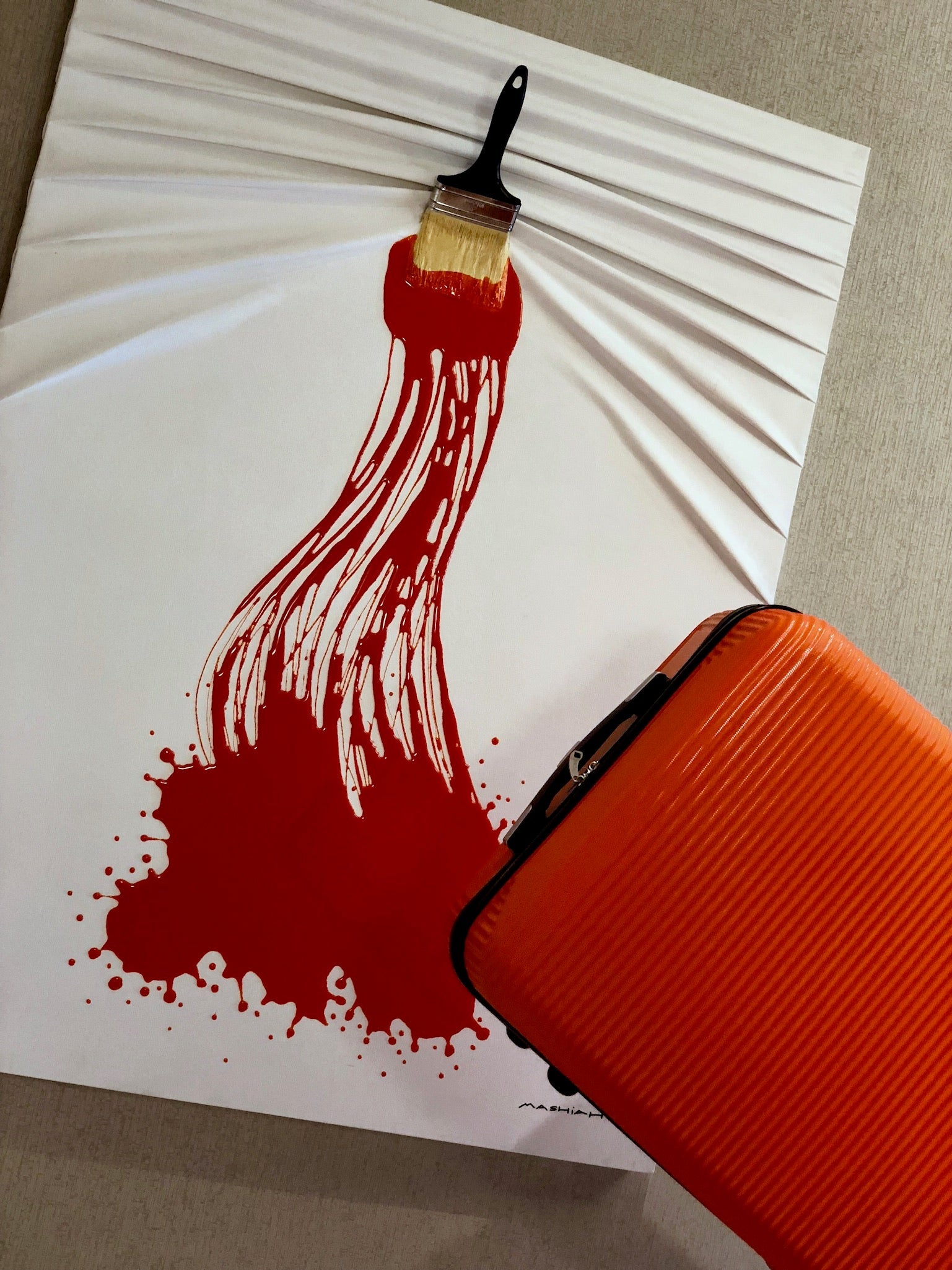 Red paint splatter with a brush on white paper, next to an orange suitcase.