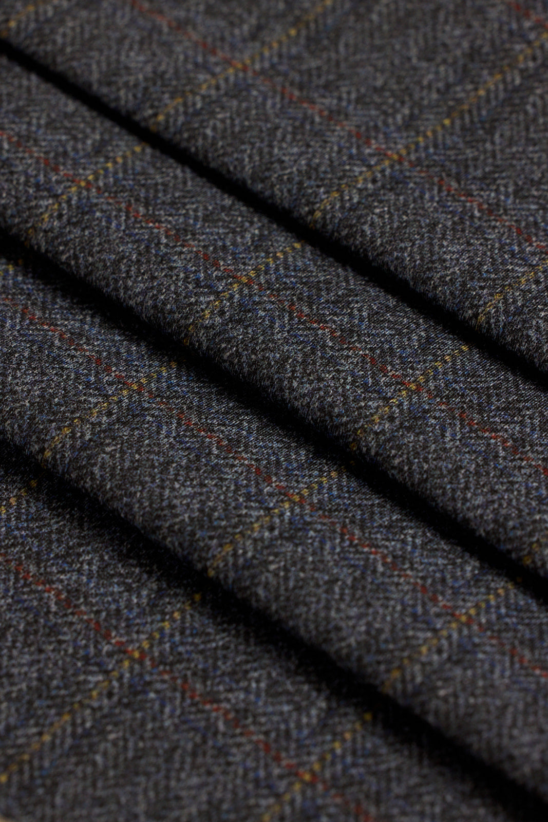 A close-up of dark gray herringbone fabric with a subtle windowpane plaid pattern in muted red, blue, and yellow lines.