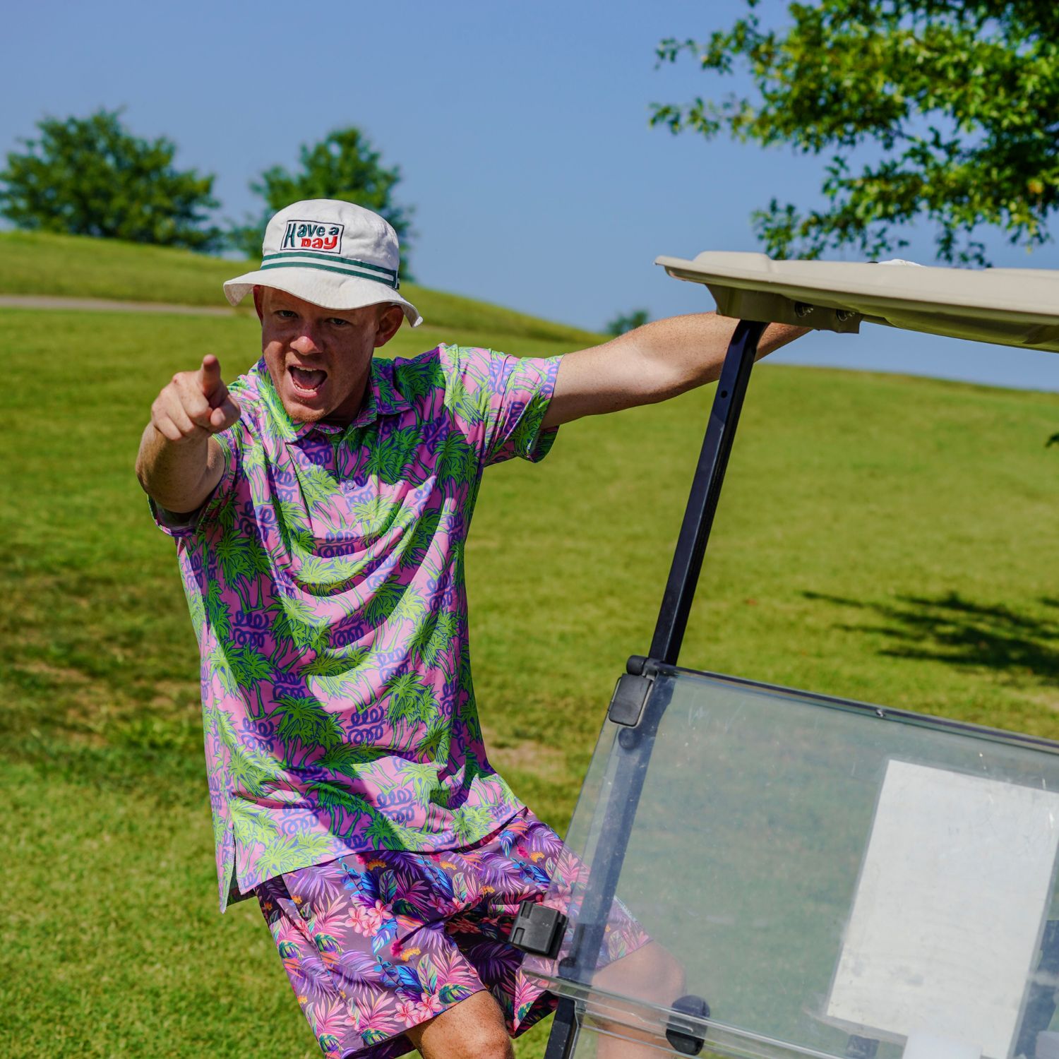 Person in colorful attire standing next to a golf cart on a grassy area with trees in the background