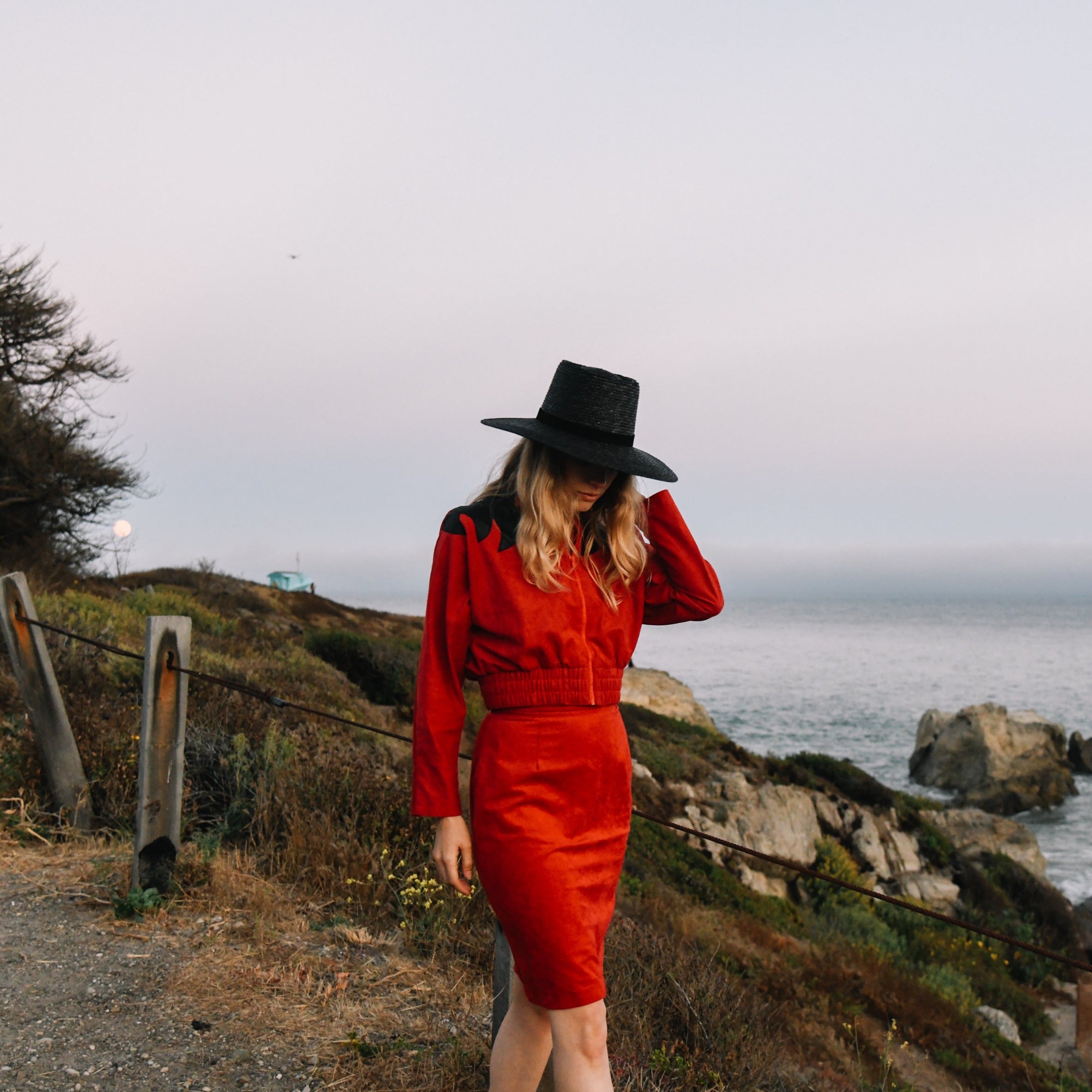Woman in a red outfit and black hat standing on a coastal path with ocean view