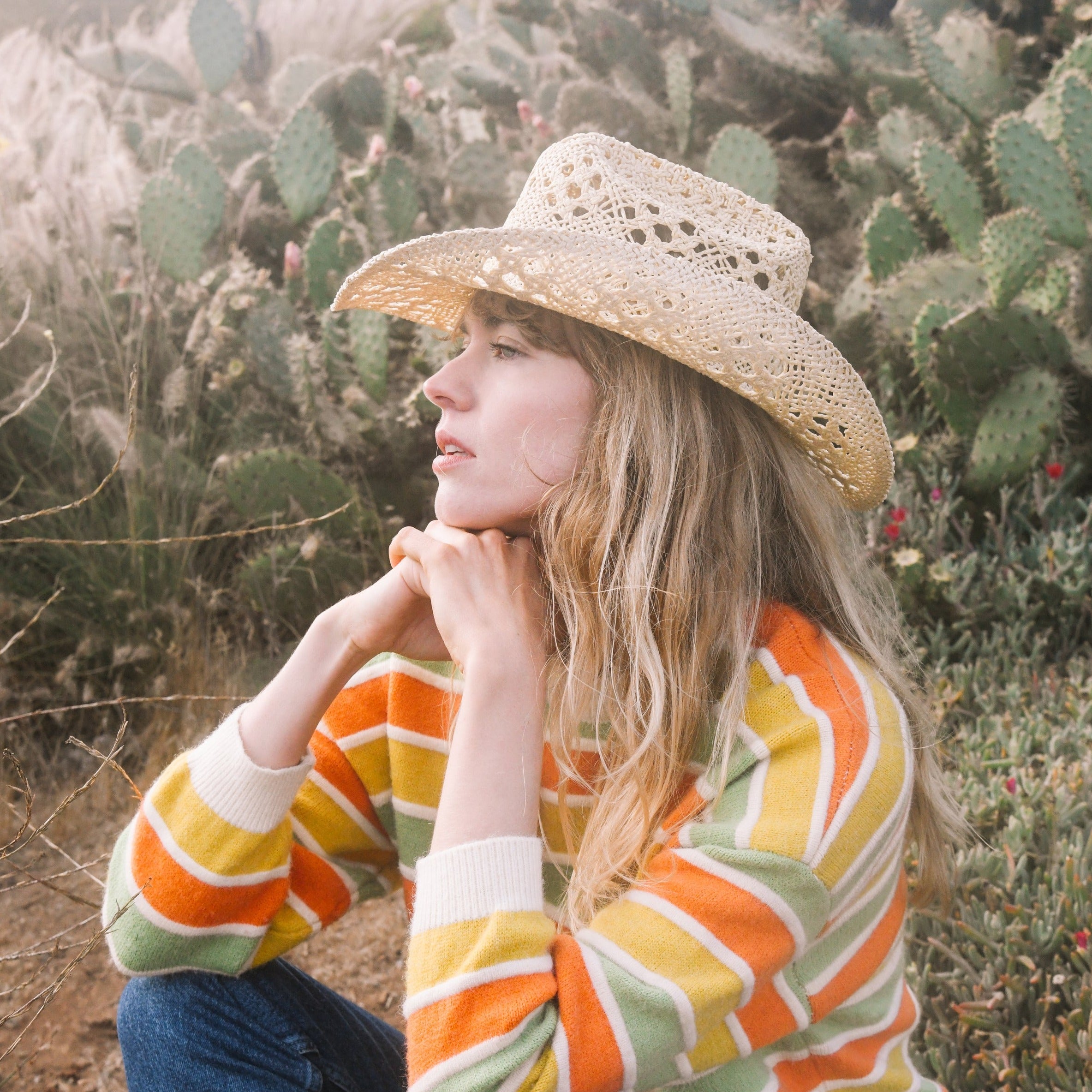 Woman wearing a straw hat and colorful striped sweater in a desert setting with cacti.