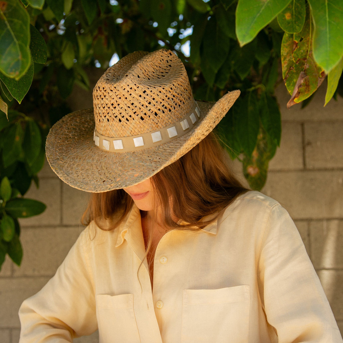 Person wearing a straw hat and light-colored shirt sitting under green leaves.