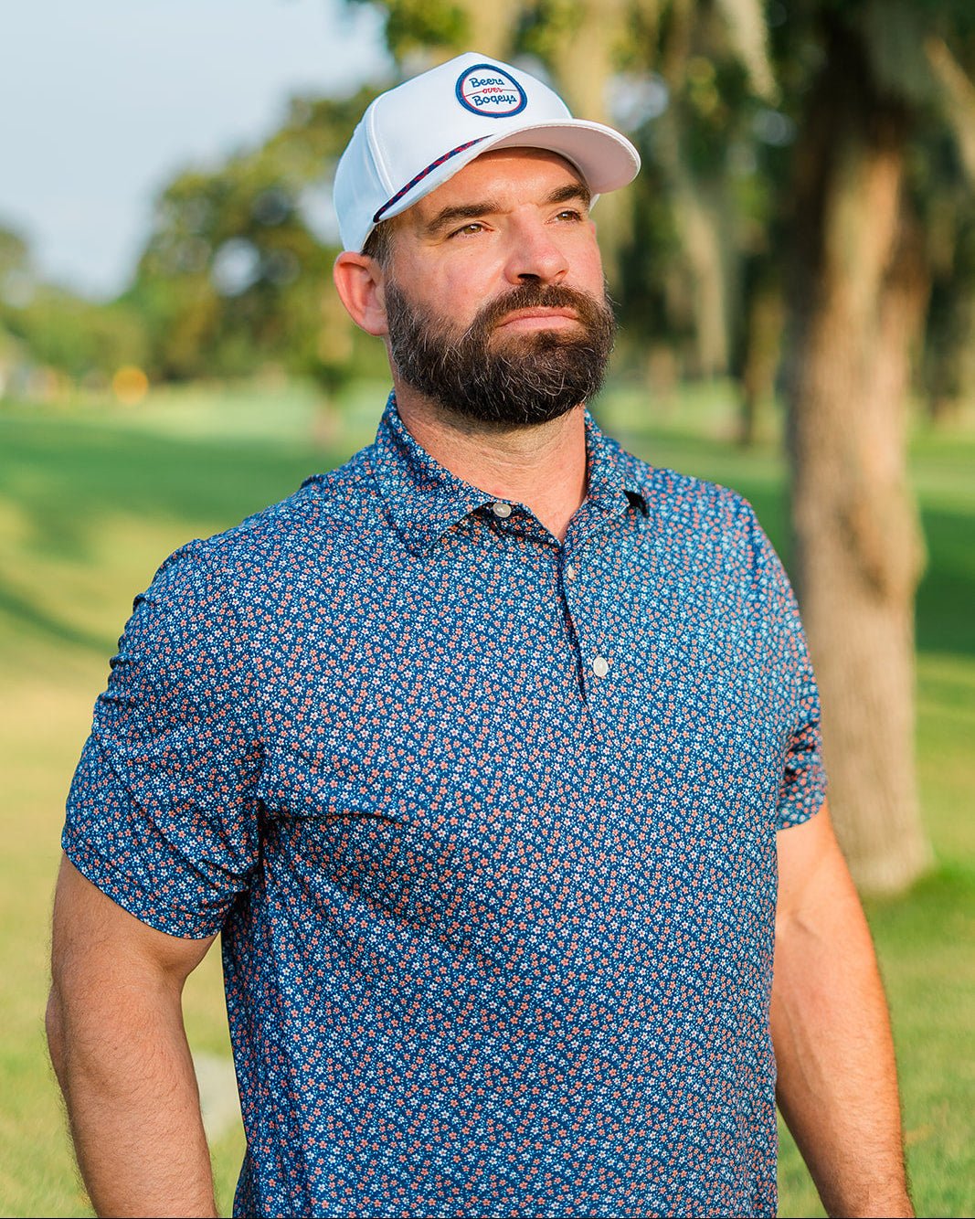 Man wearing a blue patterned shirt and white cap with a logo on a golf course