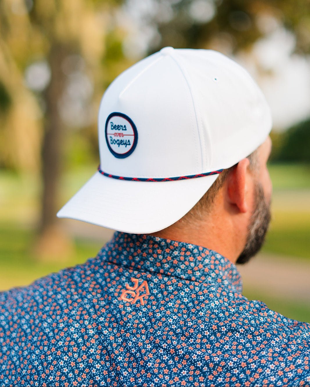 Person wearing a white cap with a logo, standing outdoors with blurred greenery in the background