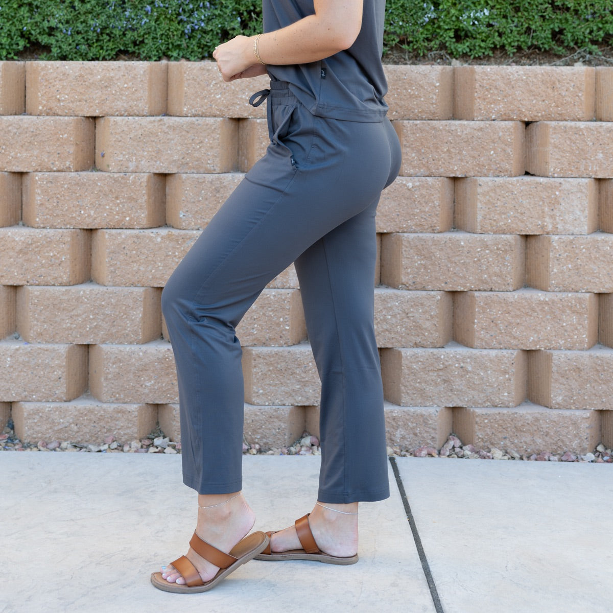 Person wearing a gray outfit with brown sandals standing in front of a stone wall.