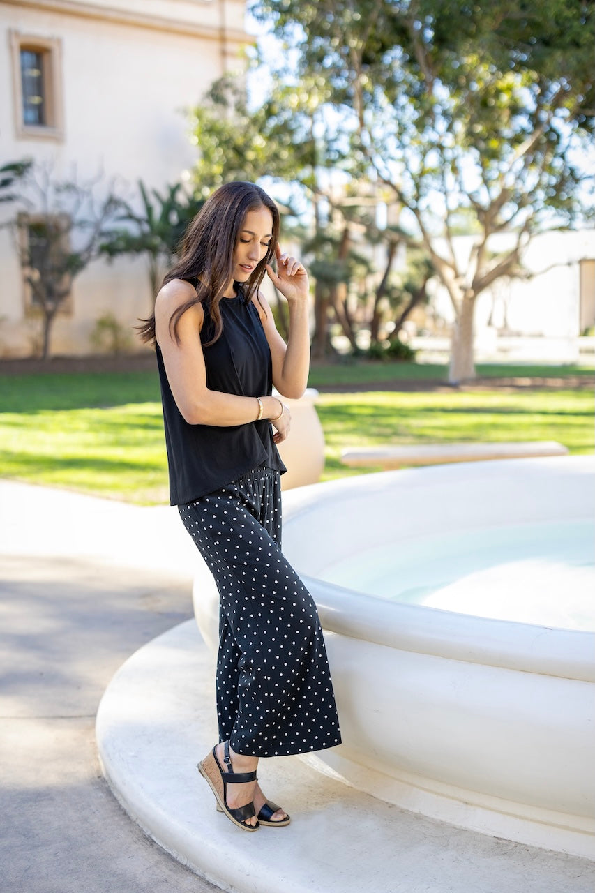 Woman in black sleeveless top and polka dot pants standing outdoors near a building with trees.