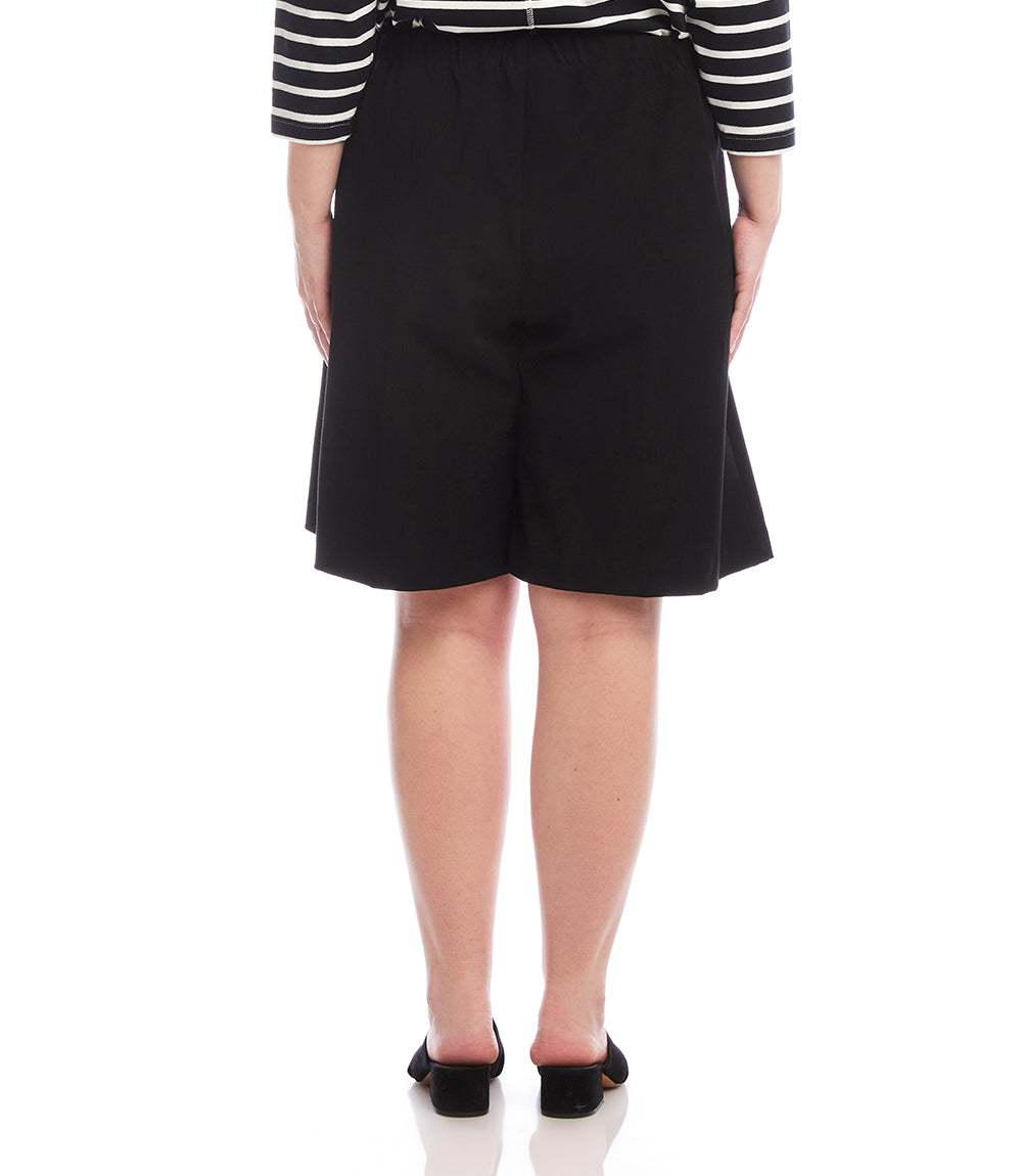 A person shown from the back wearing a black knee-length skirt or shorts-style bottom, a black-and-white striped long-sleeve top, and black flats against a plain white studio background.