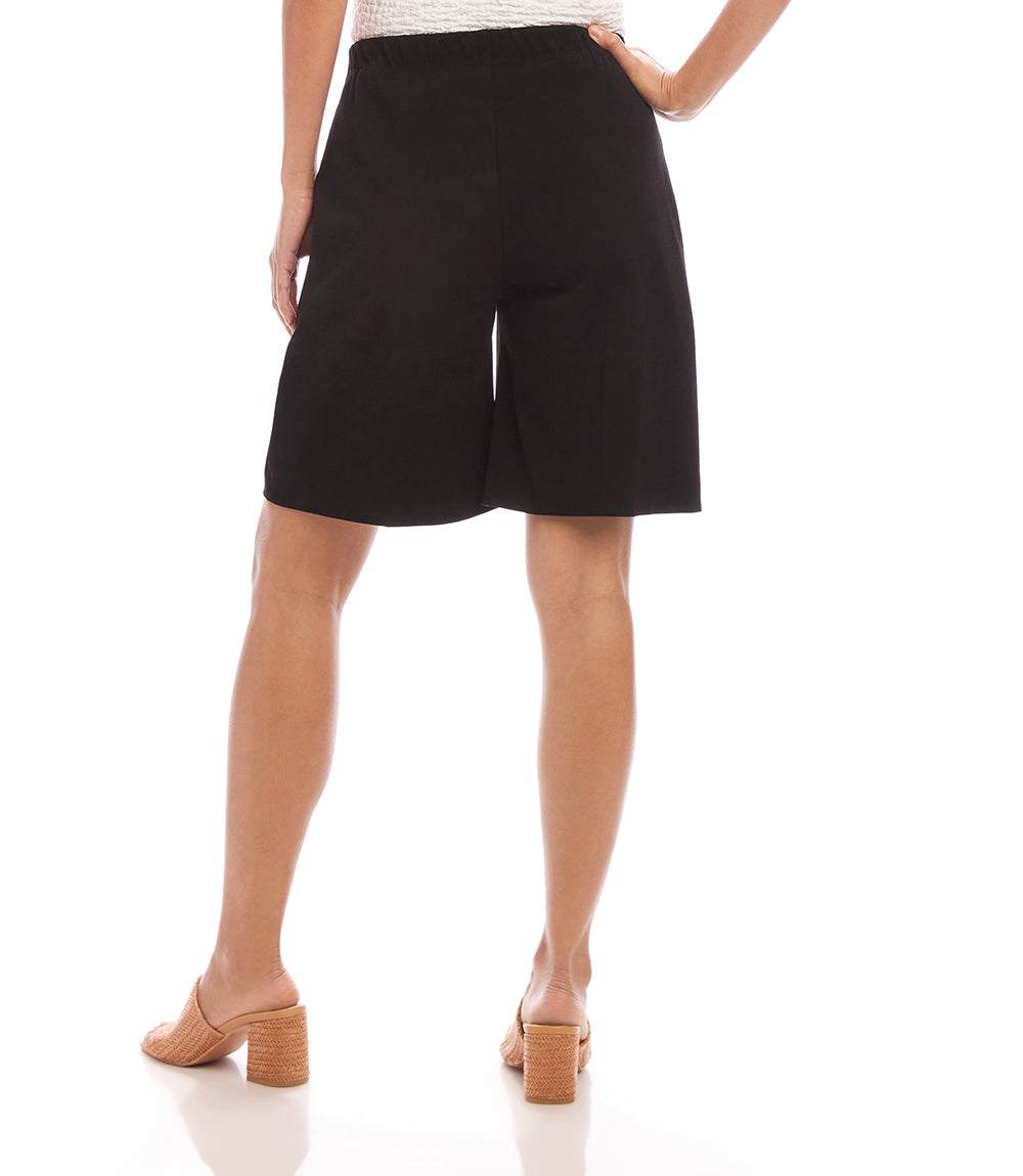 A model shown from the back wearing black wide-leg shorts, tan block-heel mule sandals, and a white top, set against a plain white studio background.