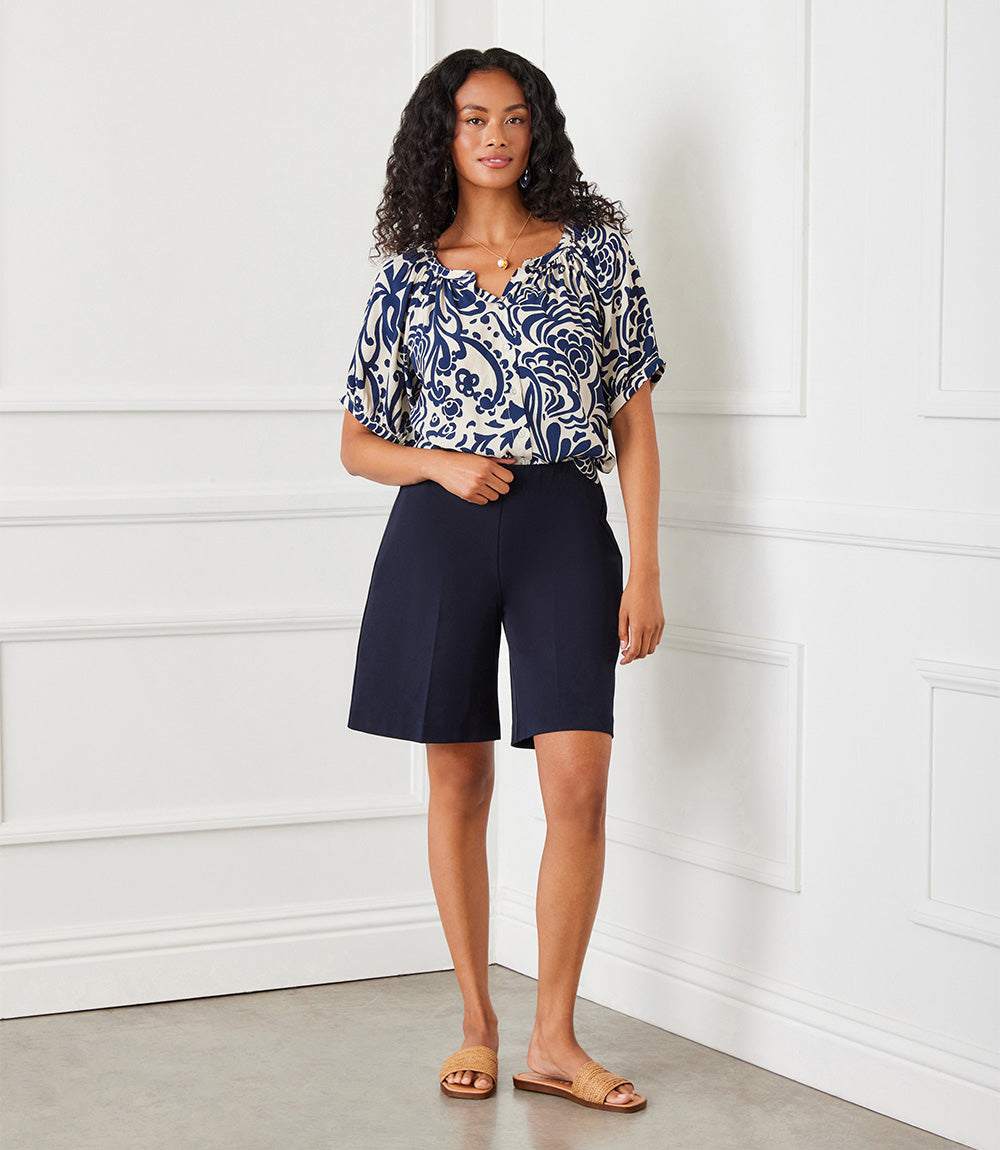 A woman stands indoors against a white paneled wall wearing a navy and white patterned short-sleeve blouse with dark navy shorts and tan slide sandals.
