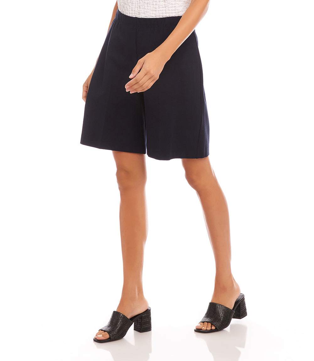 A model wearing a pair of dark navy blue high-waisted shorts, styled with a white textured top and black mule sandals, against a plain white background.