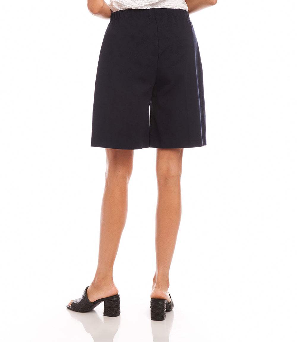 A person shown from the back wearing black high-waisted shorts with a white top and black mule sandals, photographed against a plain white studio background.